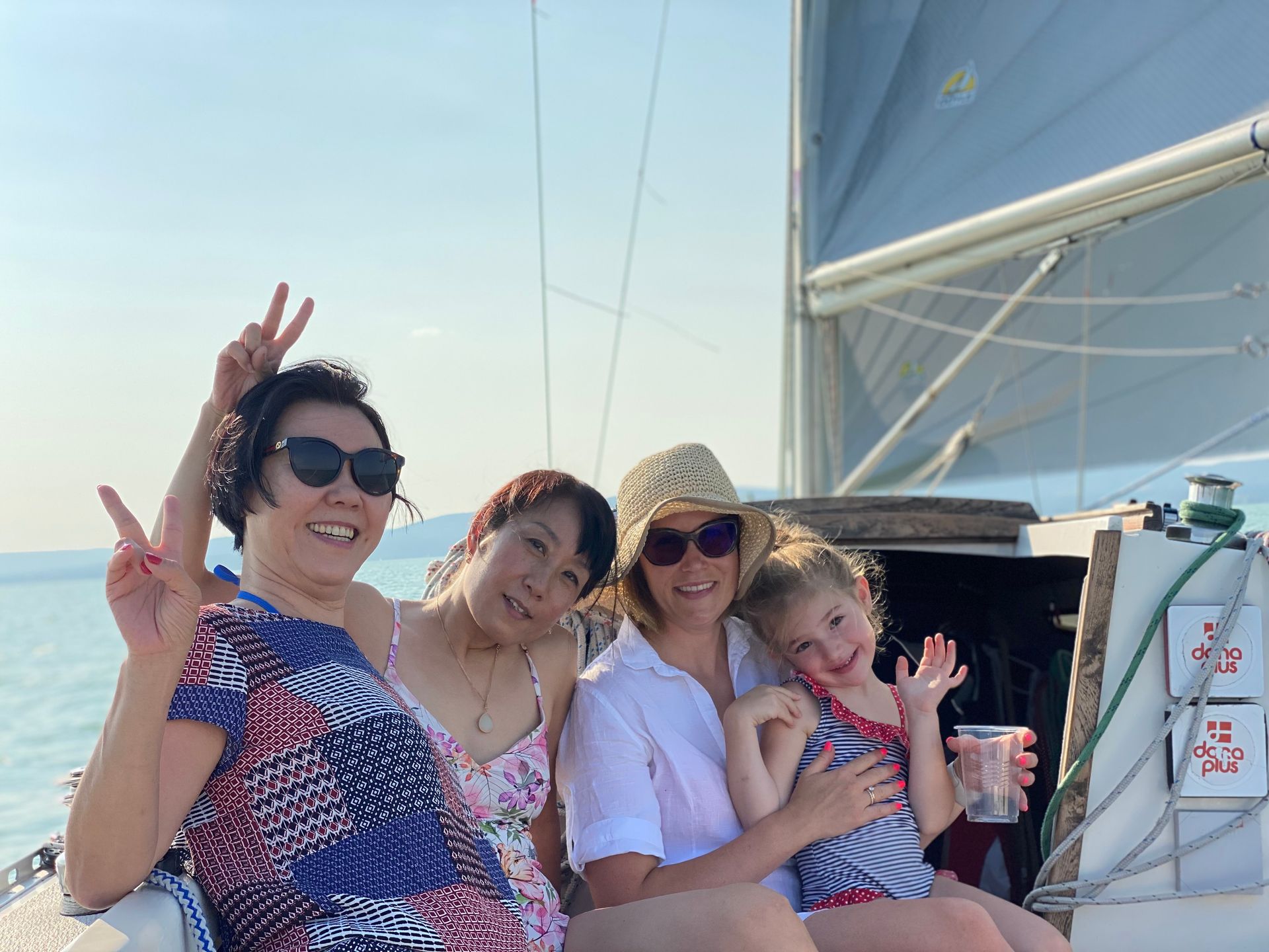 Four smiling people on a sailboat; one woman giving peace signs. Sunny day, blue water.