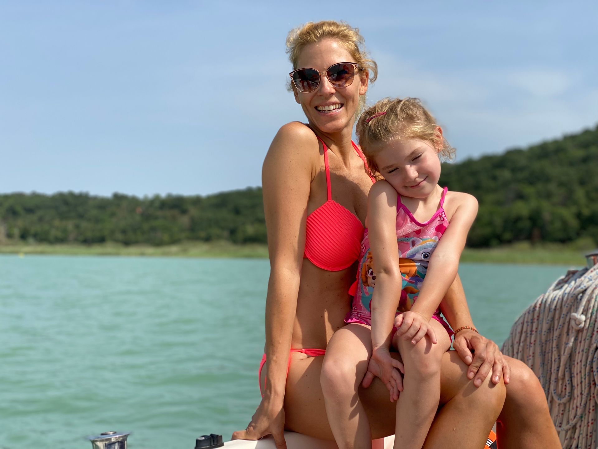 Woman in pink bikini and child in swimsuit on a boat, smiling in sunlight, overlooking water and green hills.