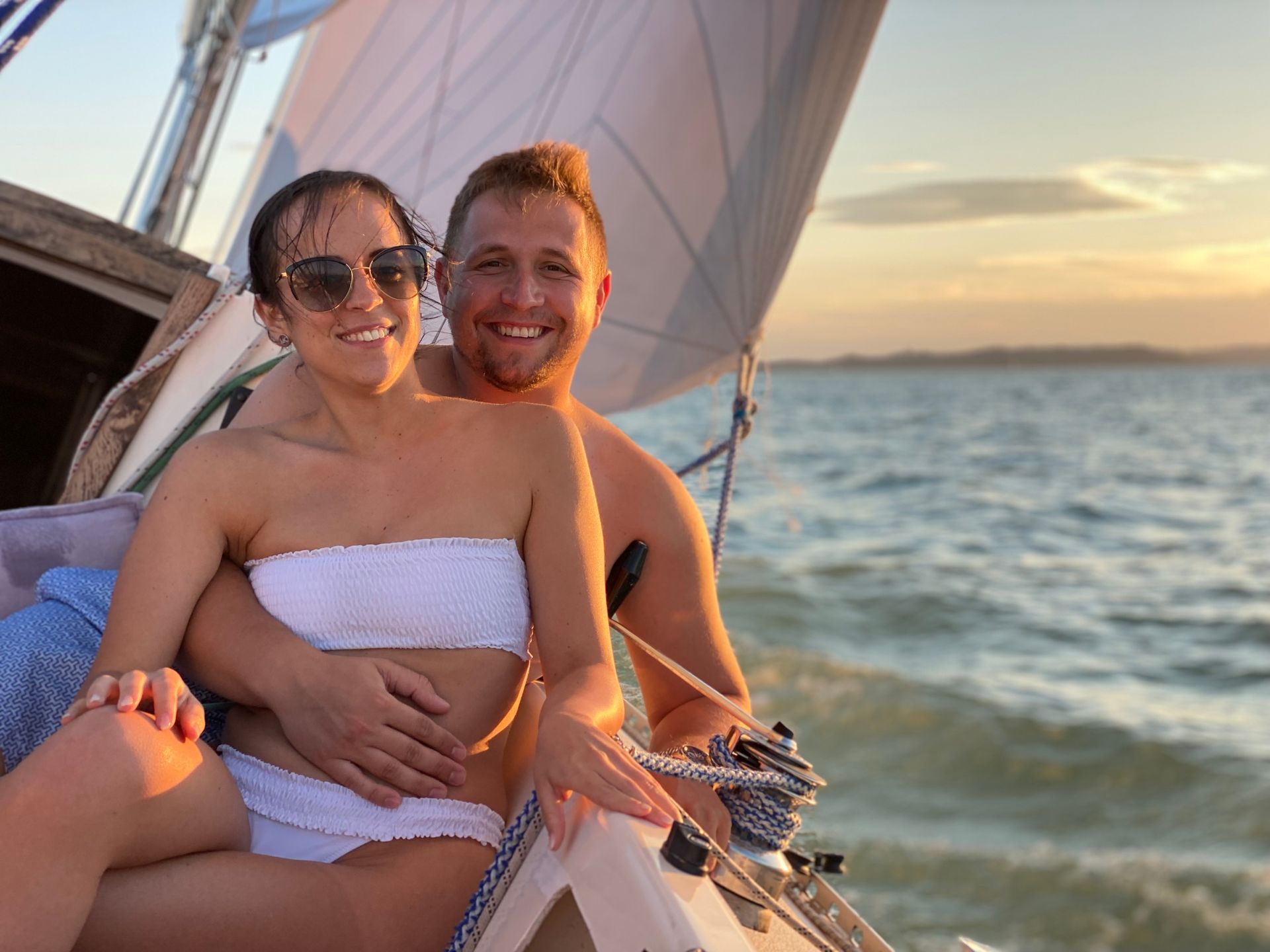Couple on a sailboat, smiling. Sunset over water in background.