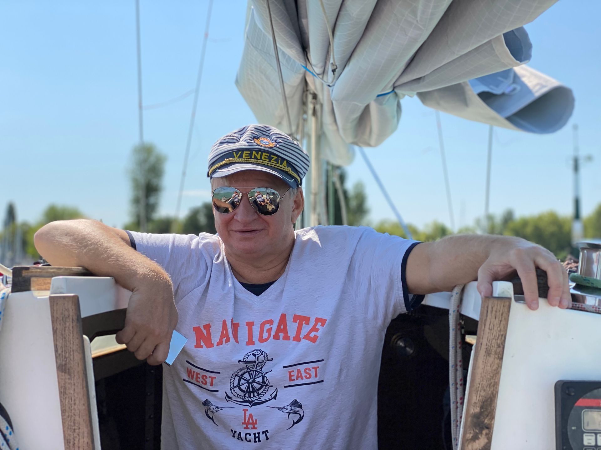Man in captain's hat and sunglasses on a sailboat, smiling. Leaning on the helm, sunny day.