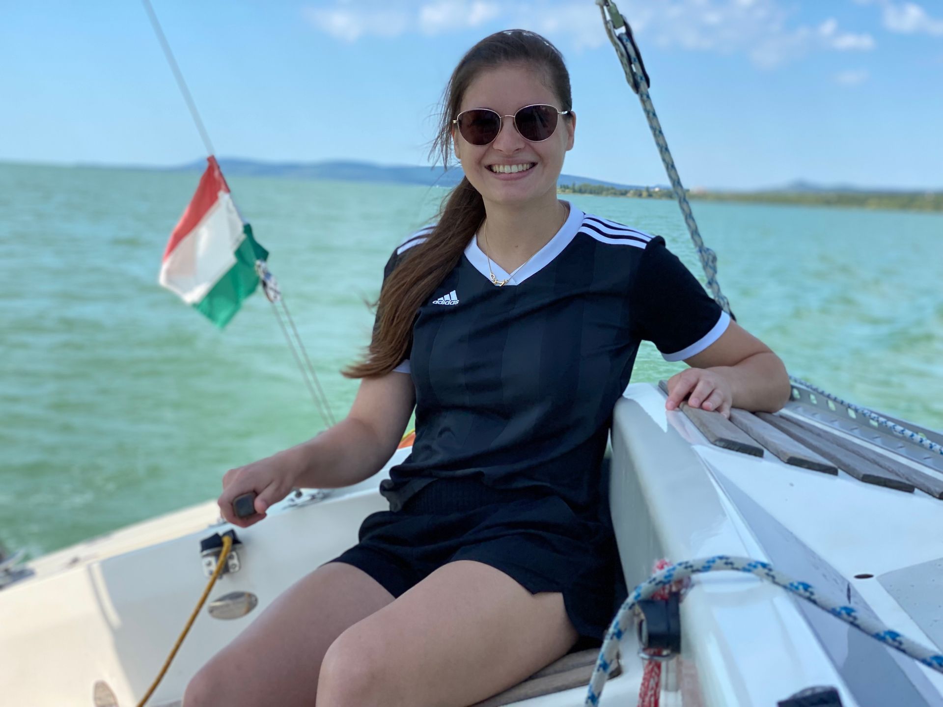 Woman sailing, smiling, wearing sunglasses and black outfit, Hungarian flag, on a boat with turquoise water.