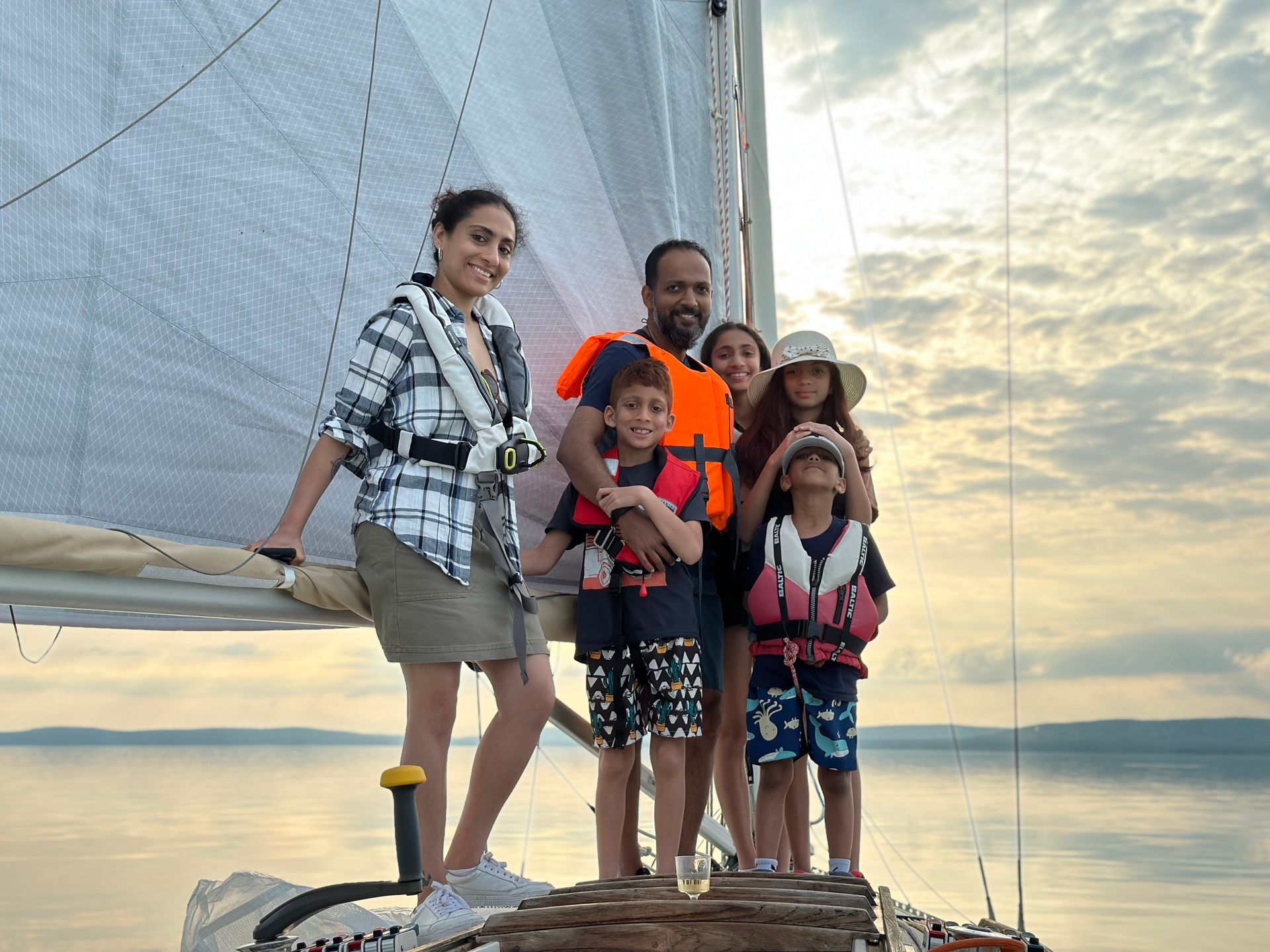 Family on sailboat, smiling. Cloudy sky, wearing life vests.