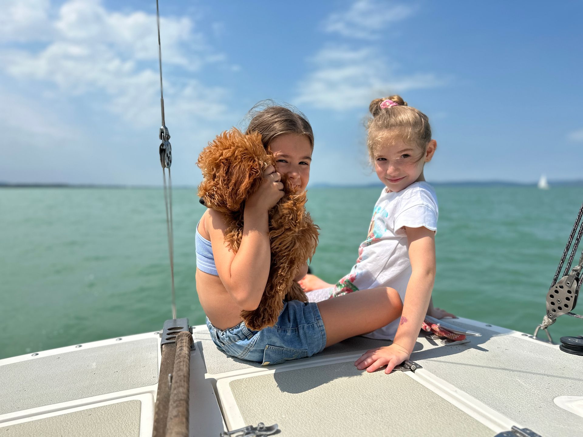 Two girls on a boat, one holding a brown dog. They smile, the sea and sky behind them.