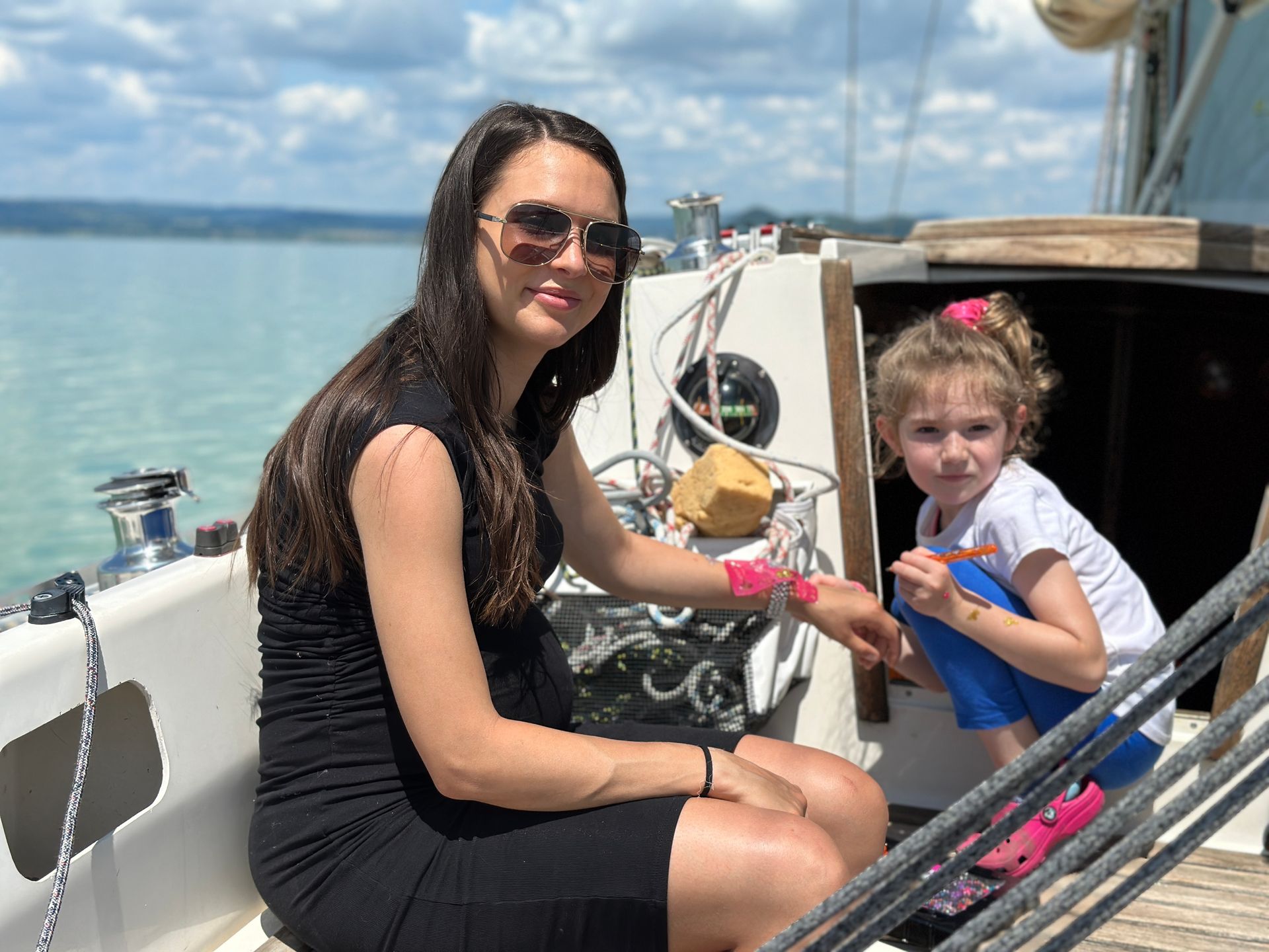 Woman and child on a sailboat. Woman in black dress and sunglasses, child in pink, blue pants. Ocean background.