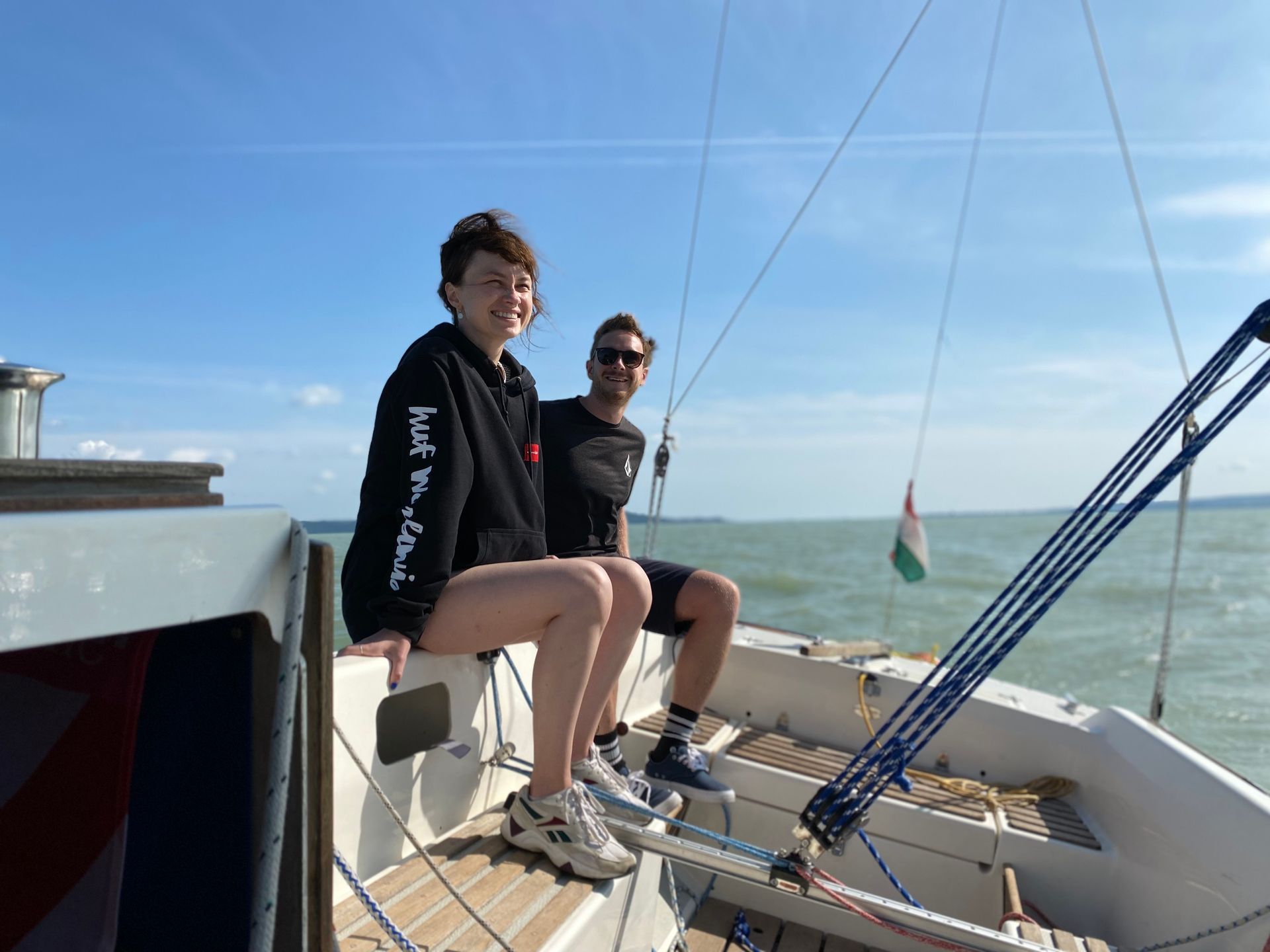 Two people on a sailboat; a smiling woman sits with legs dangling, a man smiles next to her, blue sky and water.