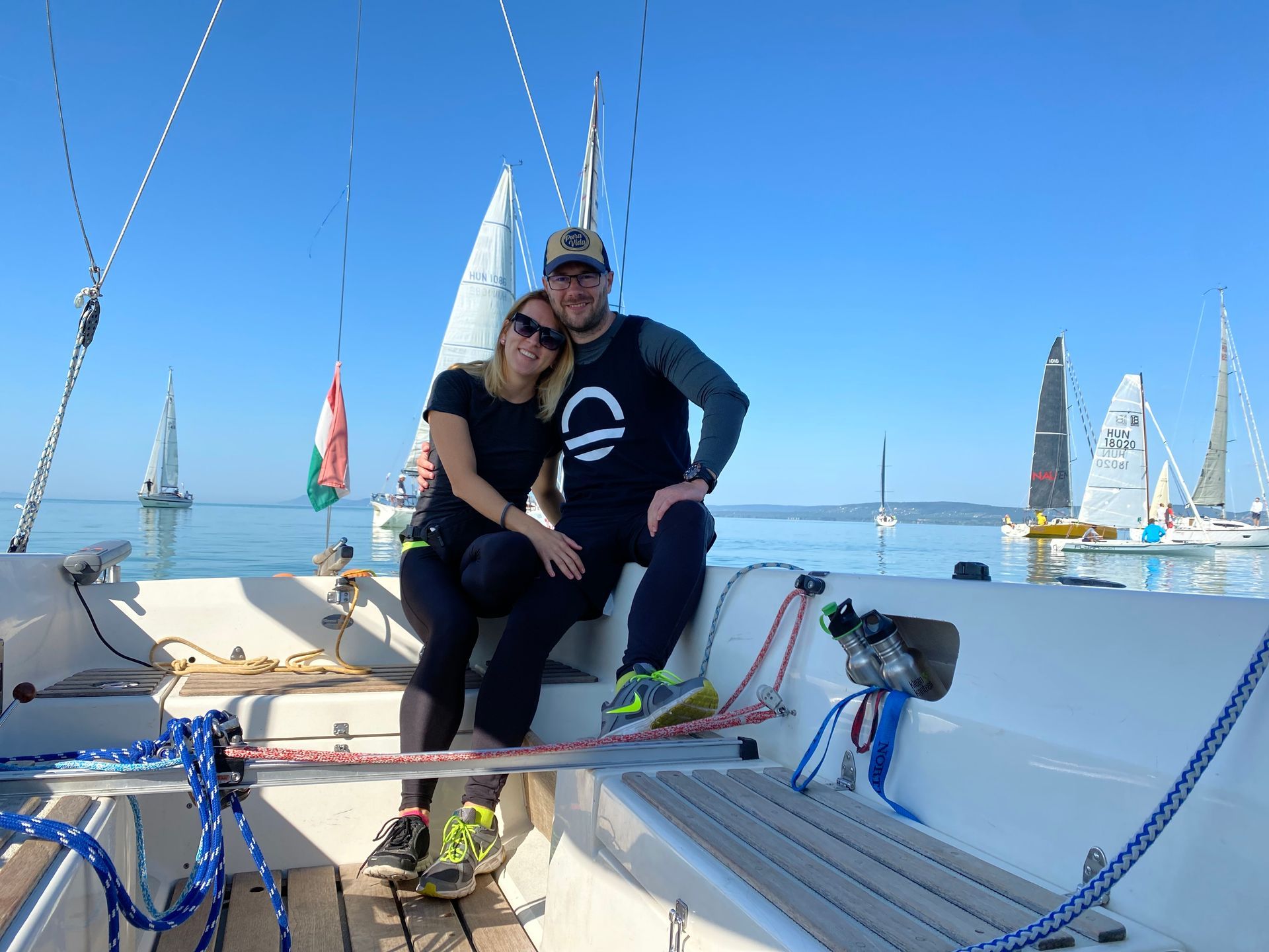 Couple on a sailboat smiling, blue water, clear sky, other sailboats in the distance.