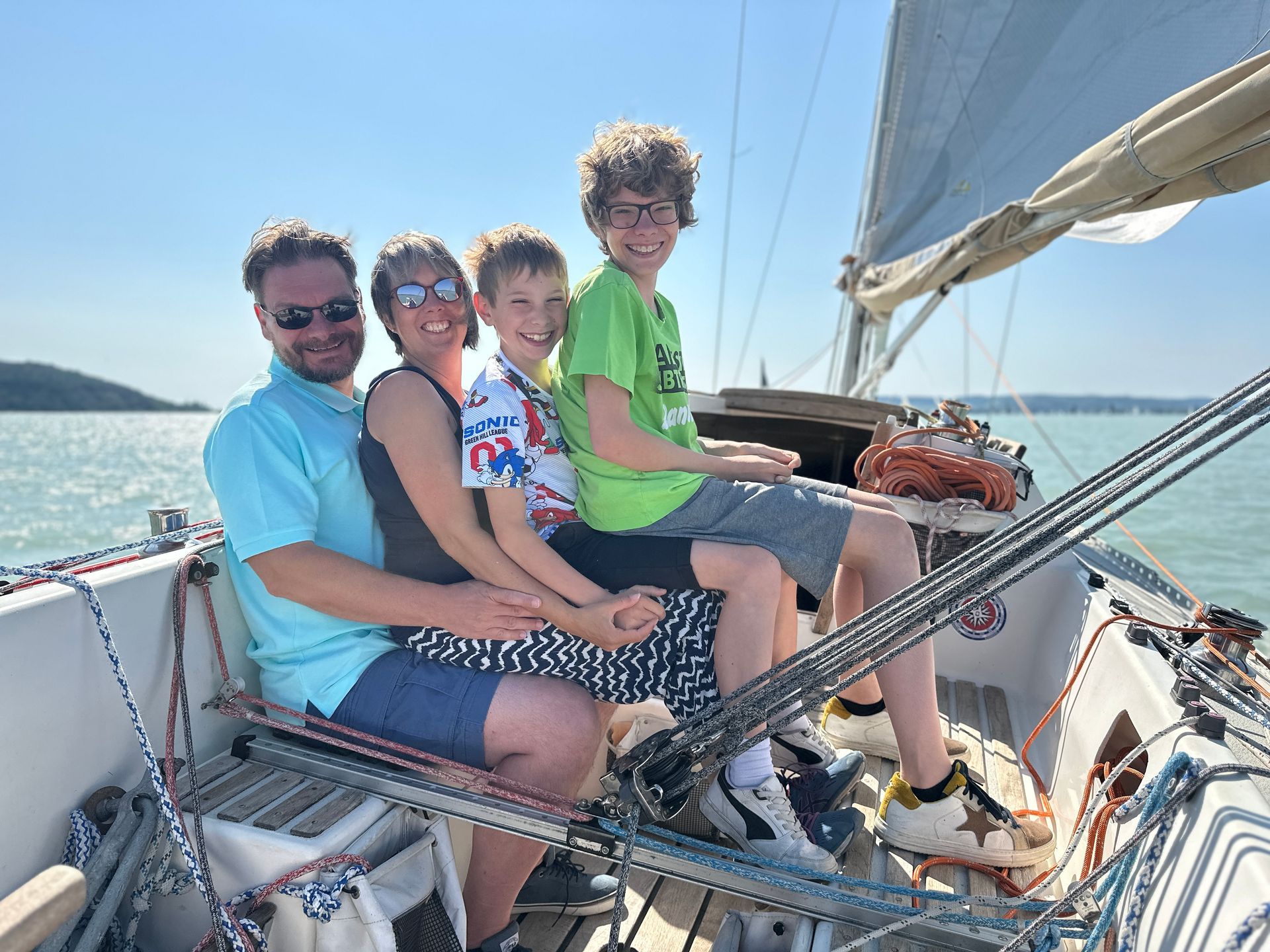 Family of four smiles on a sailboat, blue sky, turquoise water.