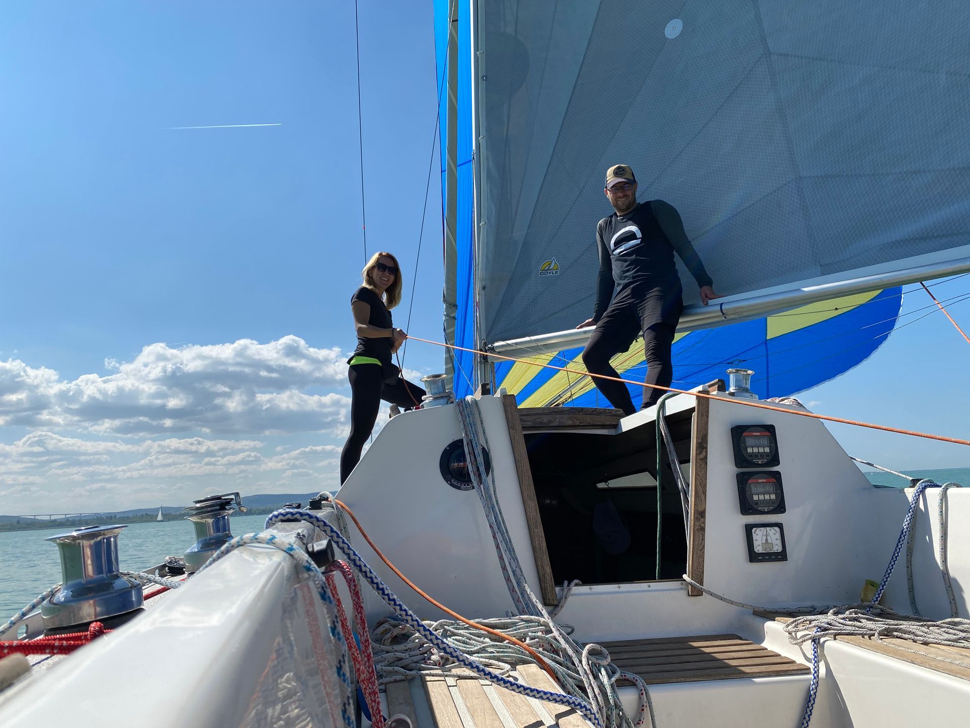 Two people on a sailboat deck under a blue sail, sunny day with coastal background.