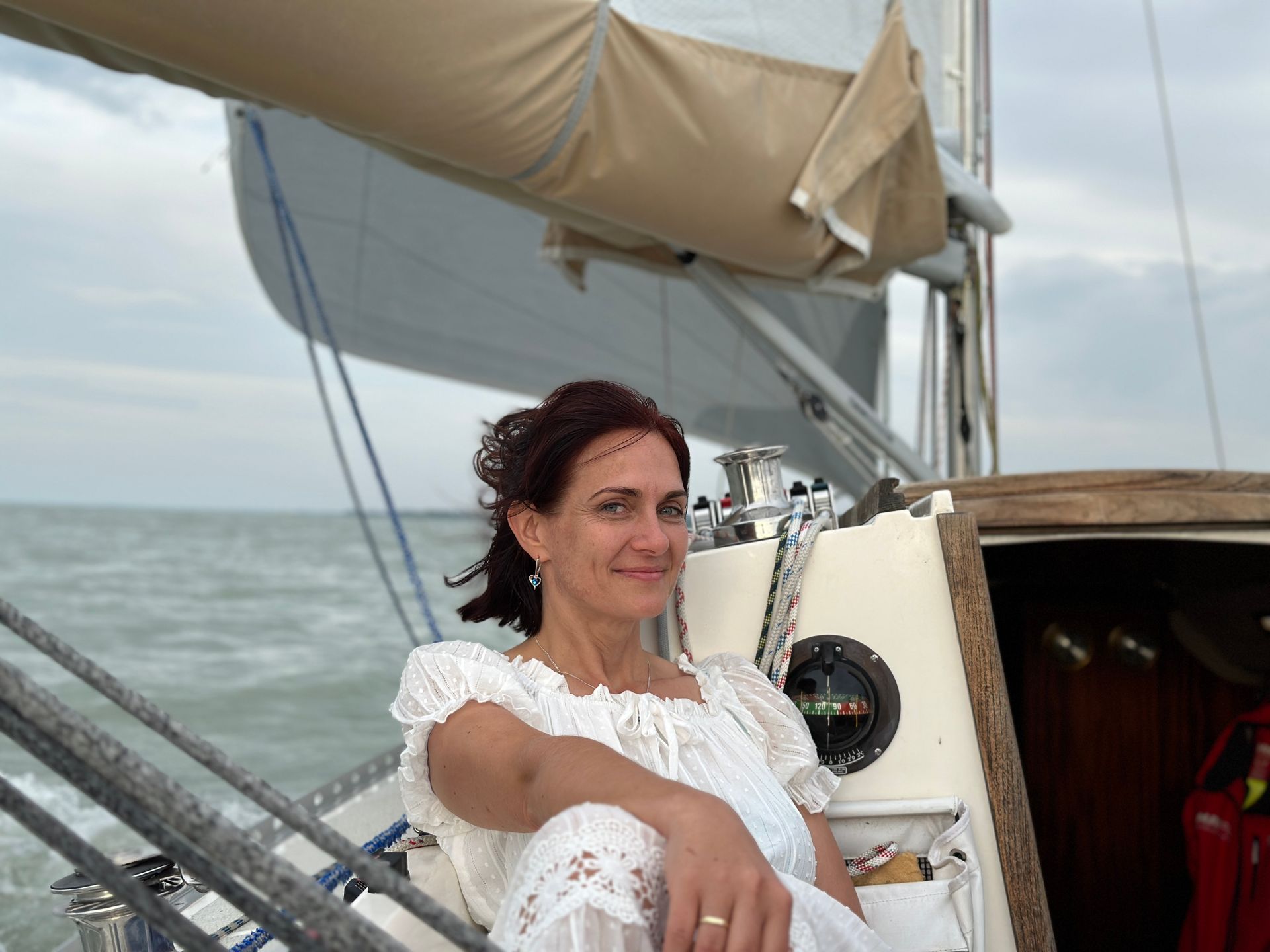 Woman with dark hair smiles on a sailboat, wearing white dress, ocean background.
