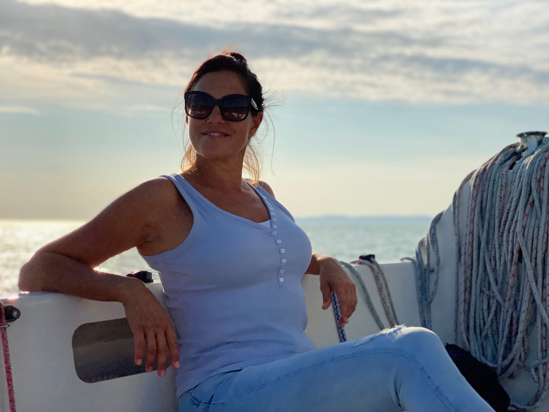 Woman wearing sunglasses and a tank top, seated on a sailboat, smiles at the sea.