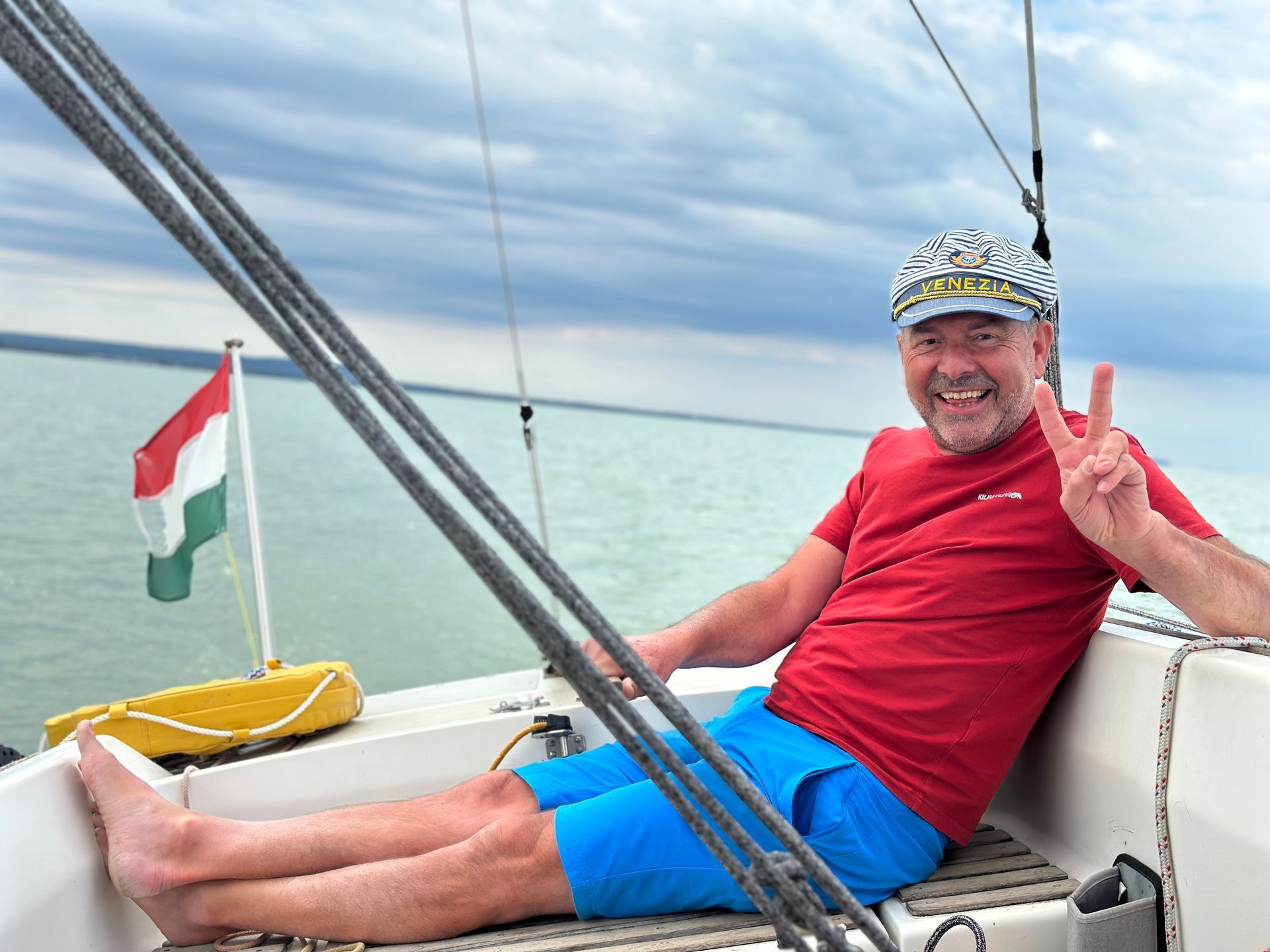 Man relaxing on a sailboat, wearing a red shirt and blue shorts, smiling and holding up a peace sign.