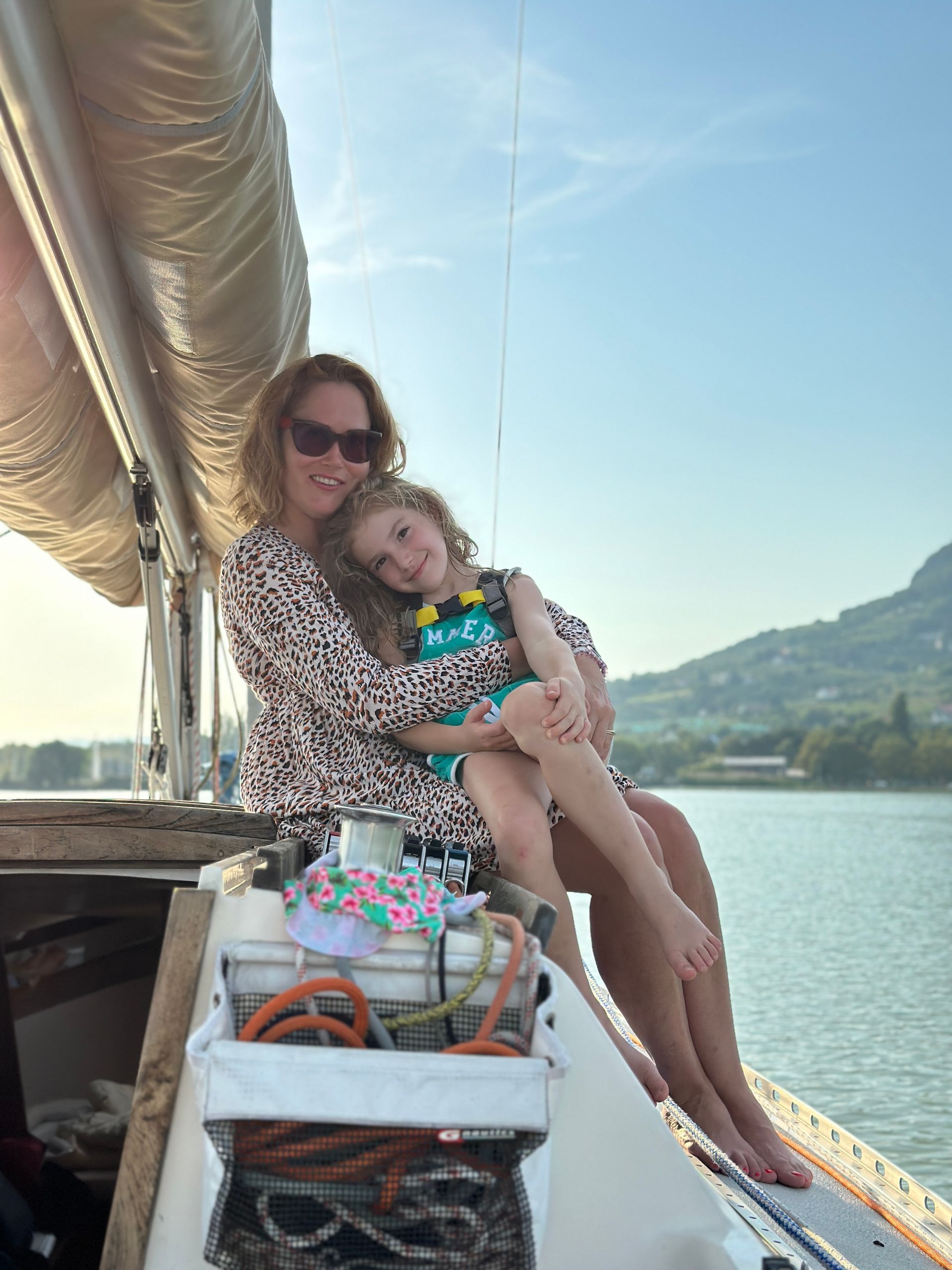 Woman and child on a sailboat, posing for a photo. Sky, water, and a small mountain in the background.