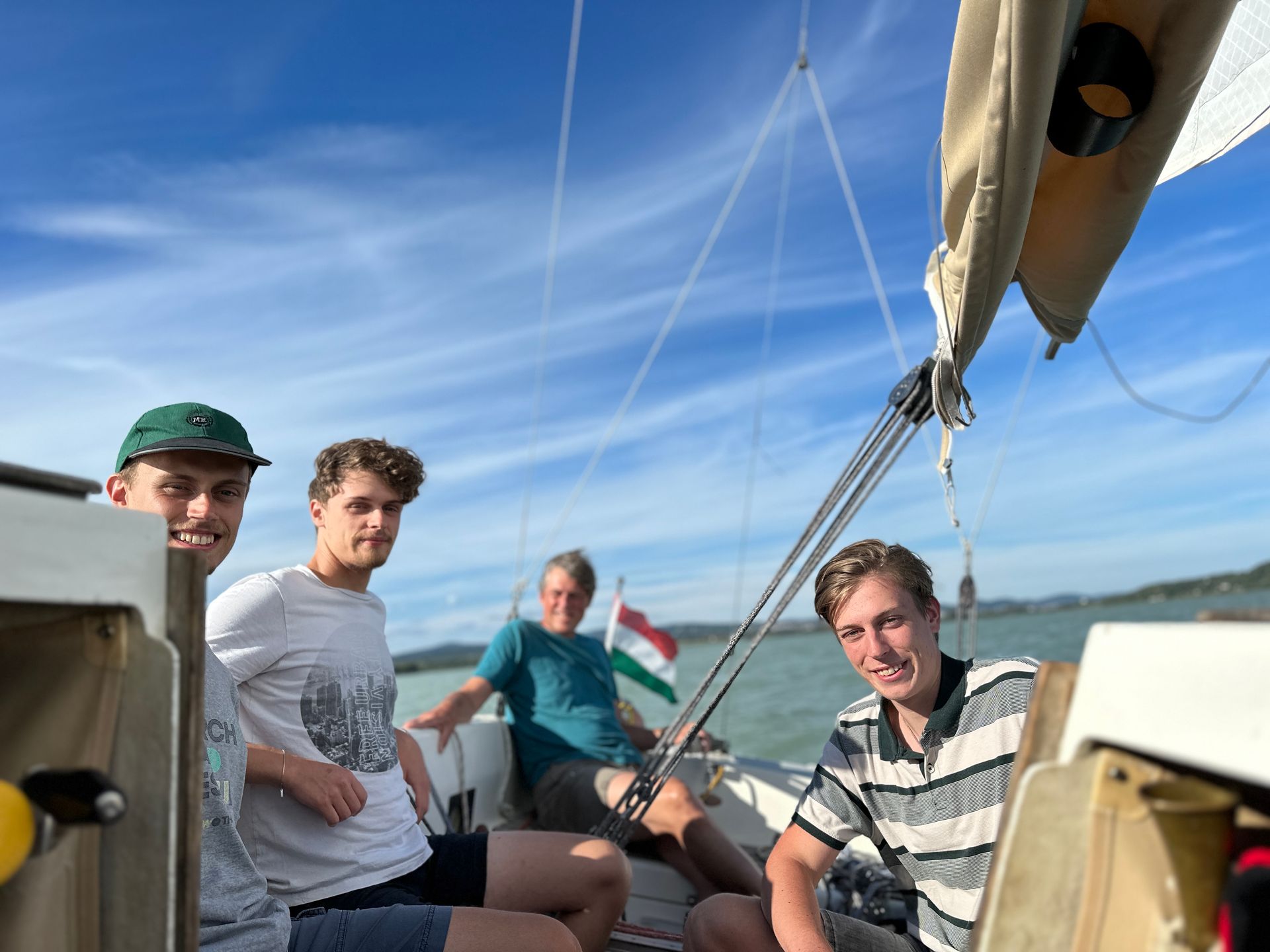 Four young men sailing on a boat under a blue sky; one holds a sail rope, Hungary flag.