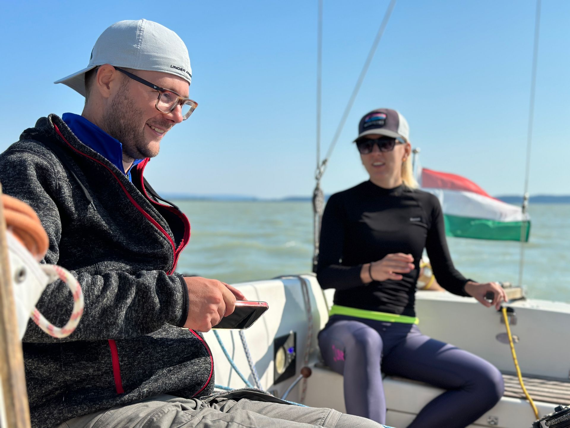 Man using phone smiles on a sailboat with woman and Hungarian flag in background.