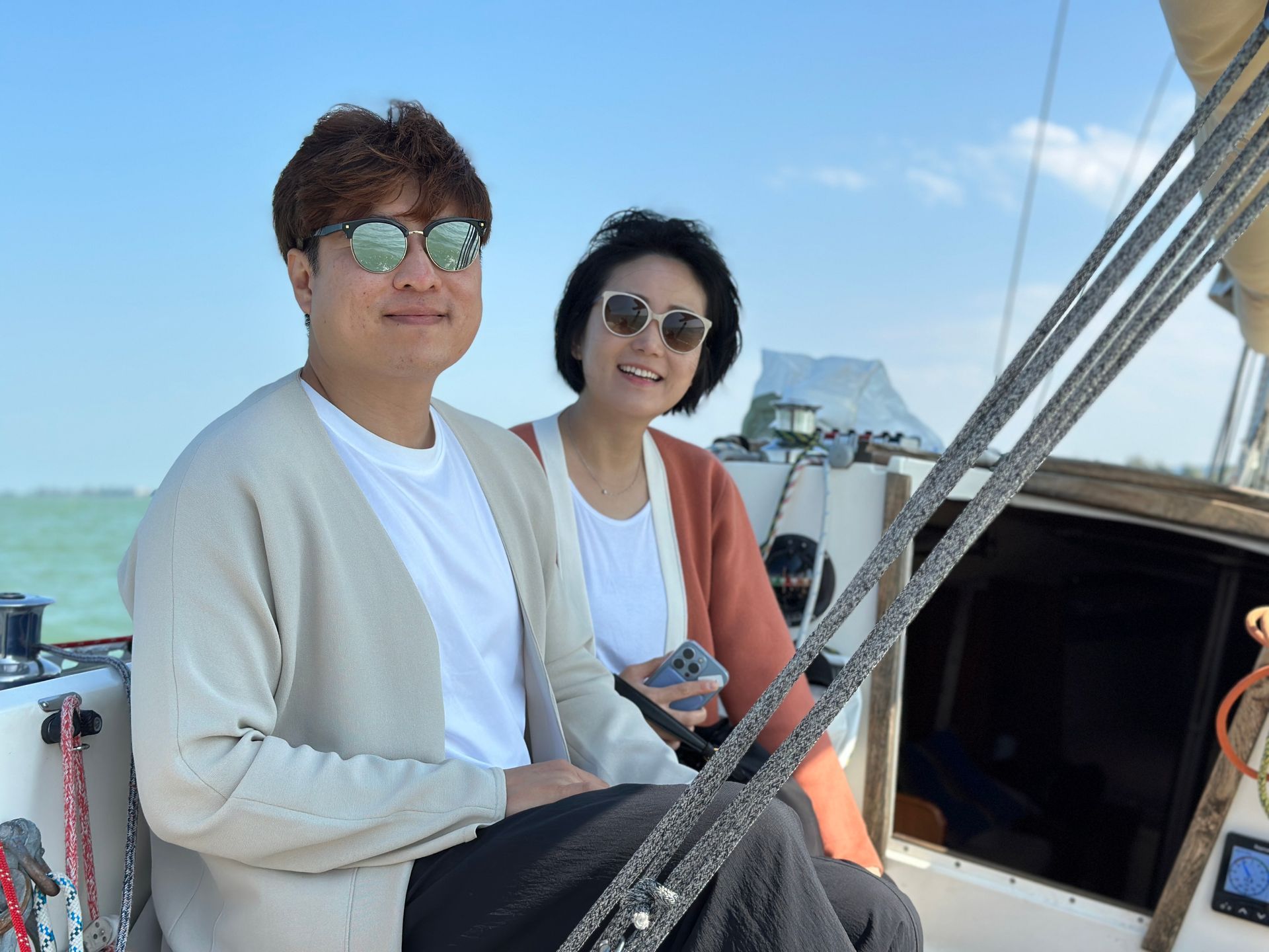 Two people on a sailboat, smiling at the camera. Sunny day, clear blue sky.