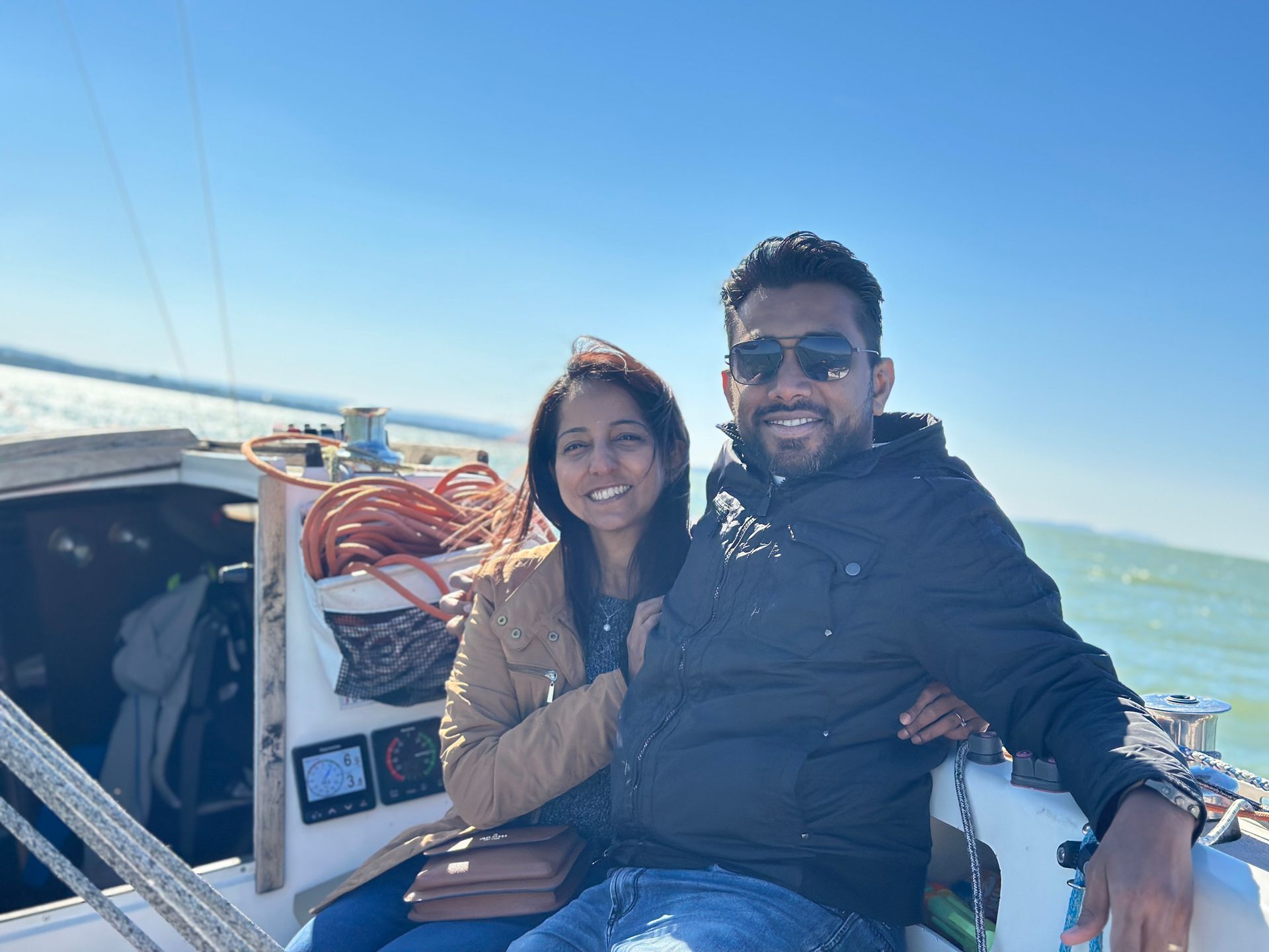 Two people smiling on a boat in bright sunlight, with blue water and sky behind them