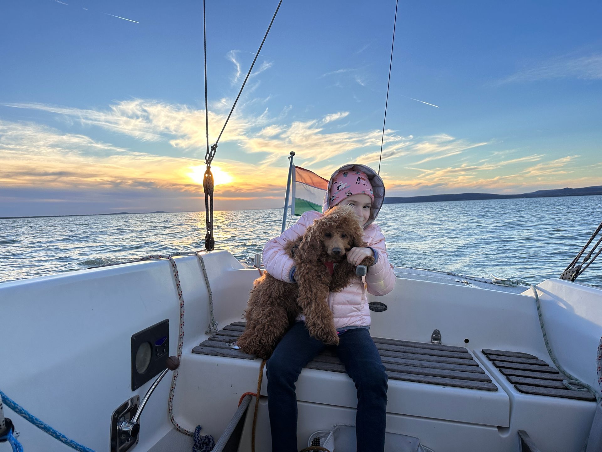 Girl in pink jacket holding a brown dog on a sailboat, with a sunset and water.