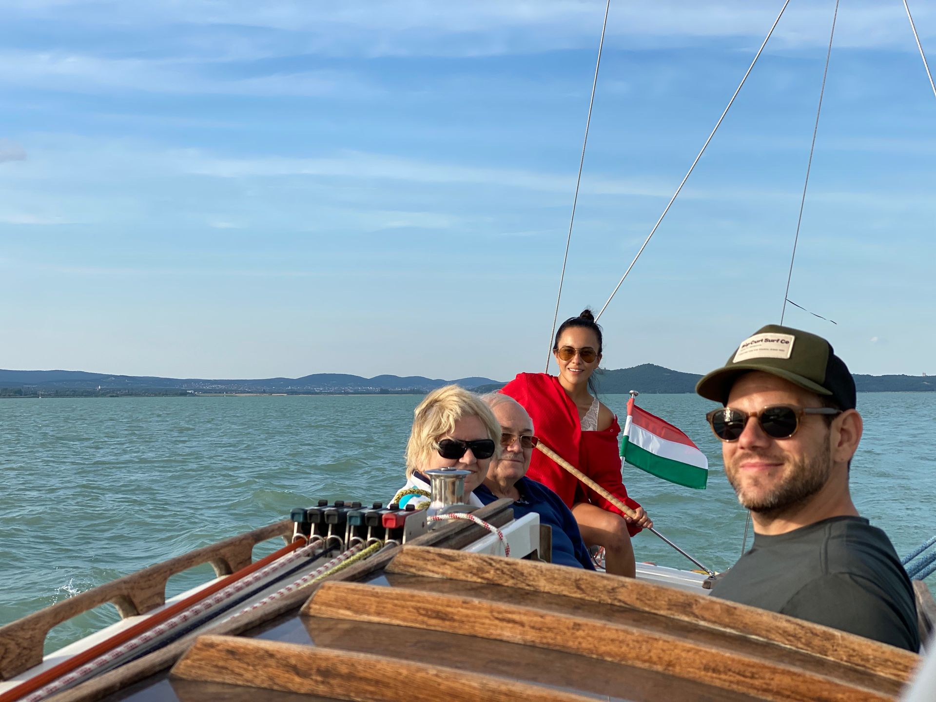 People on a sailboat with a Hungarian flag. Blue water and sky. Smiling faces, sunny day.