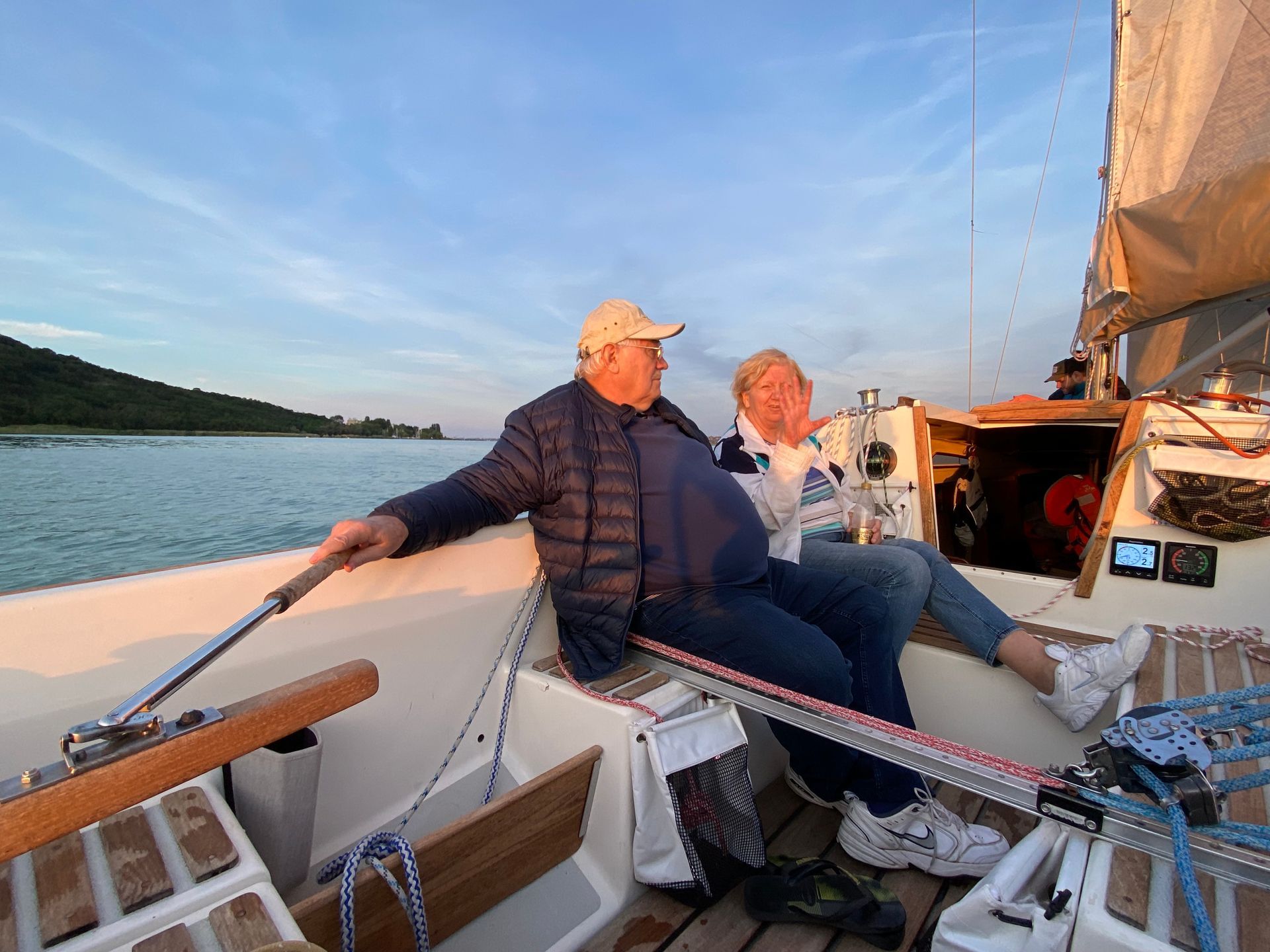Two people on a sailboat, smiling, enjoying a sunset. Man steering, woman holding drink.