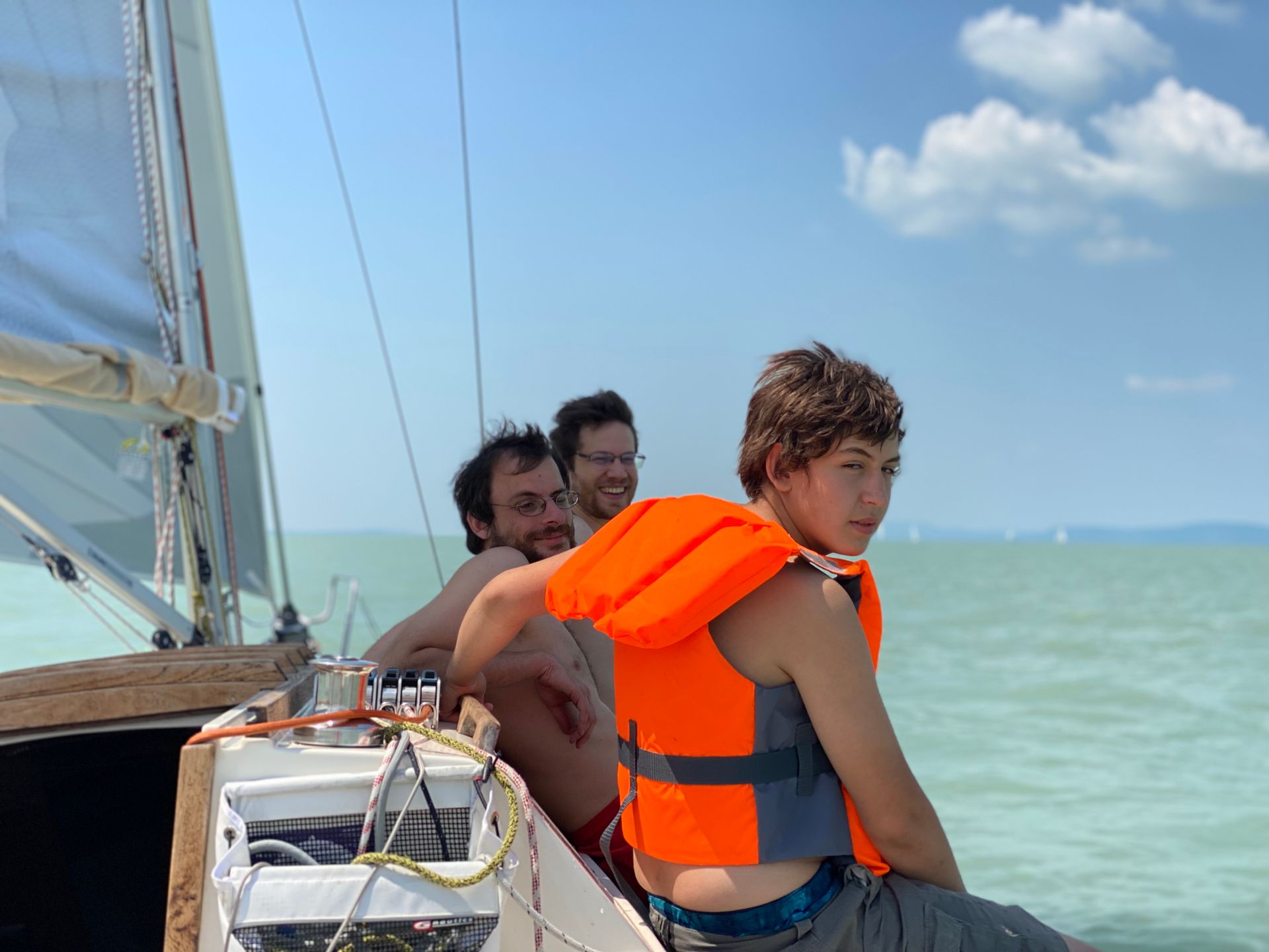 Three people on a sailboat, two smiling, one wearing an orange life vest, sailing on a sunny day.