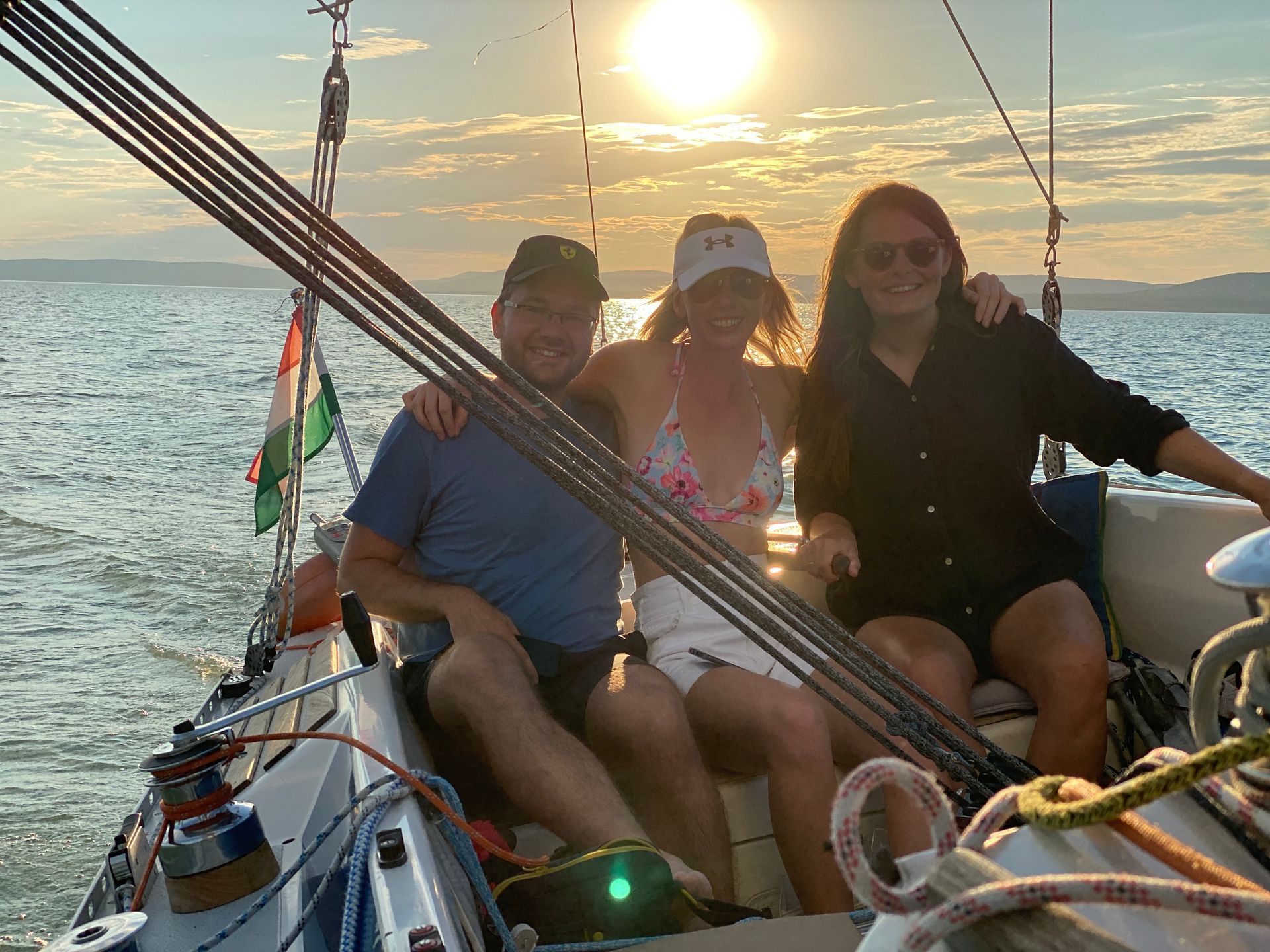 Three people smiling on a sailboat at sunset. The sun is setting over the water.