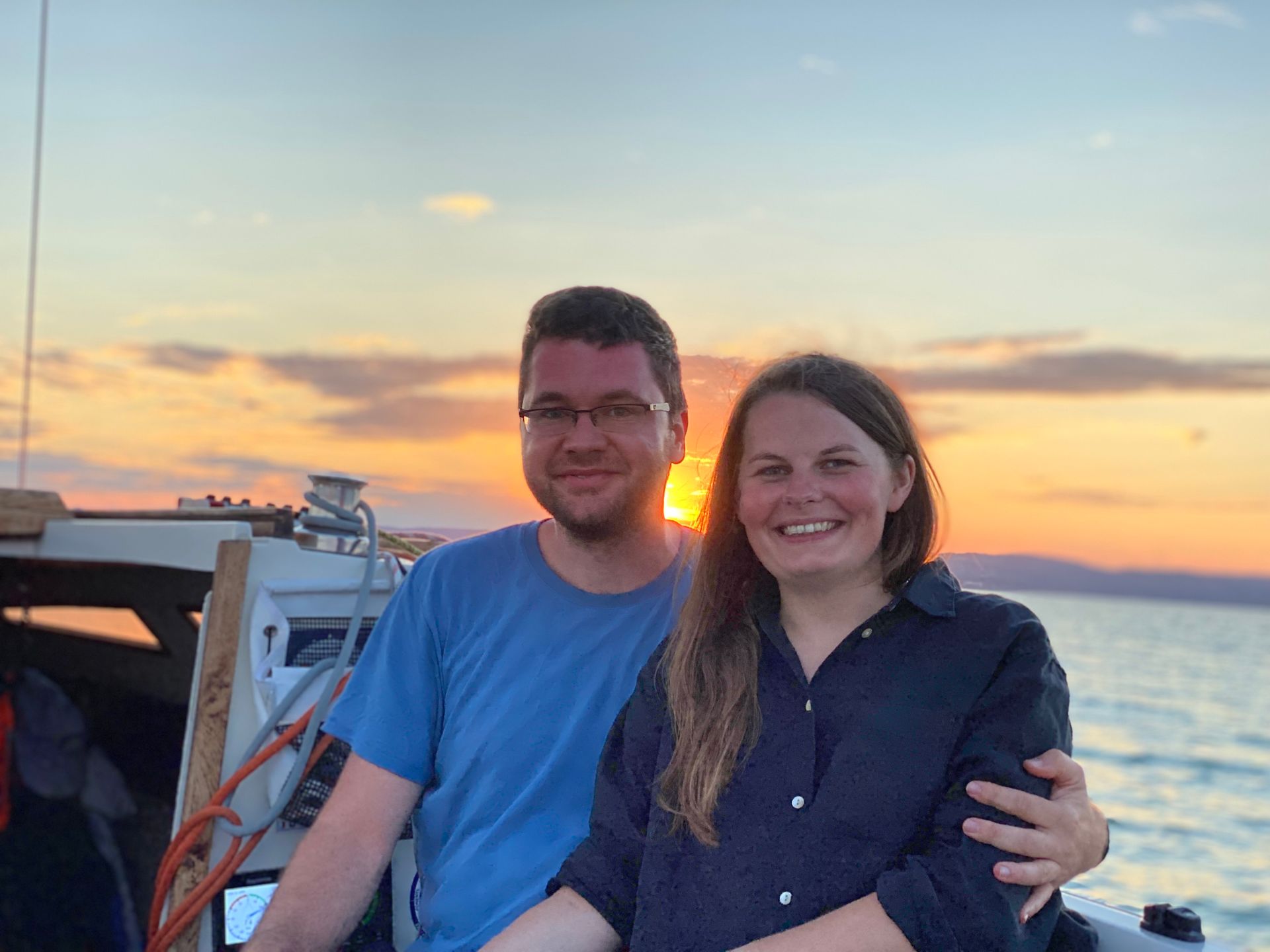 Man and woman on a boat at sunset, embracing. The sky is orange and the water is calm.