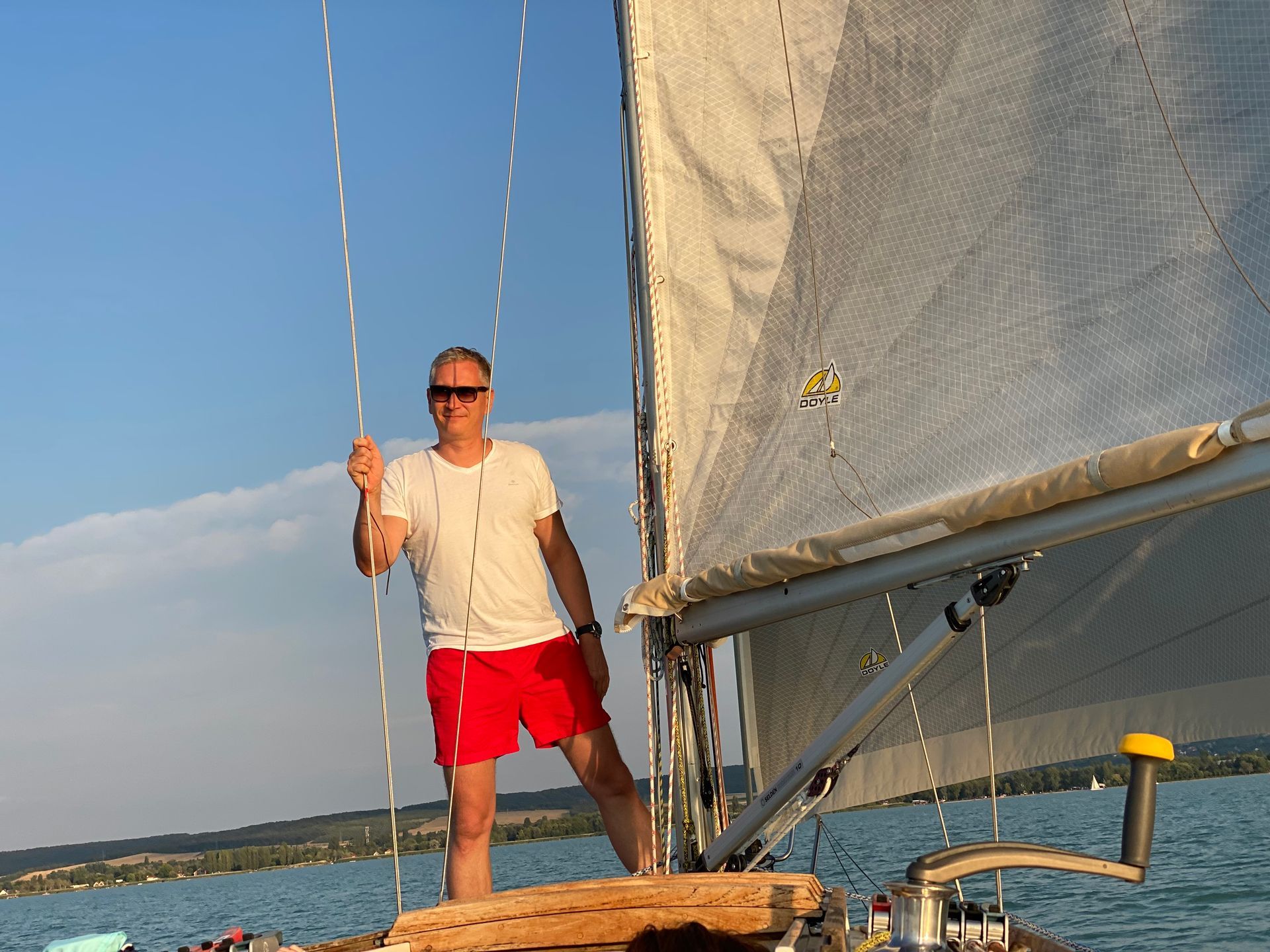 Man in red shorts sails a boat, holding a rope; blue sky and water.