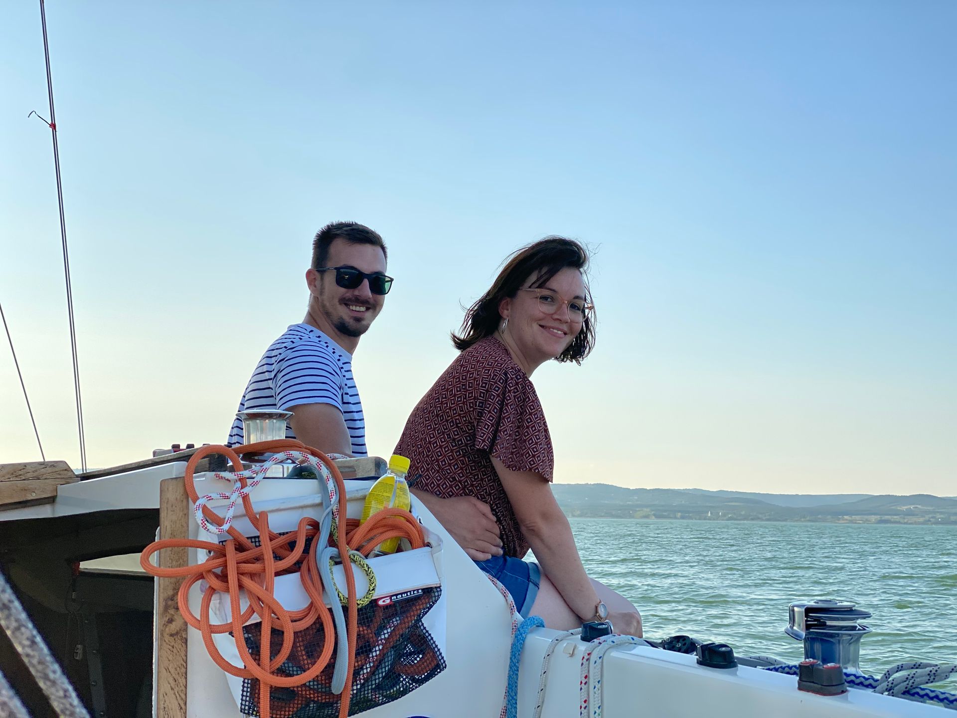 A man and woman smile while seated on a boat, with blue water and sky in the background.