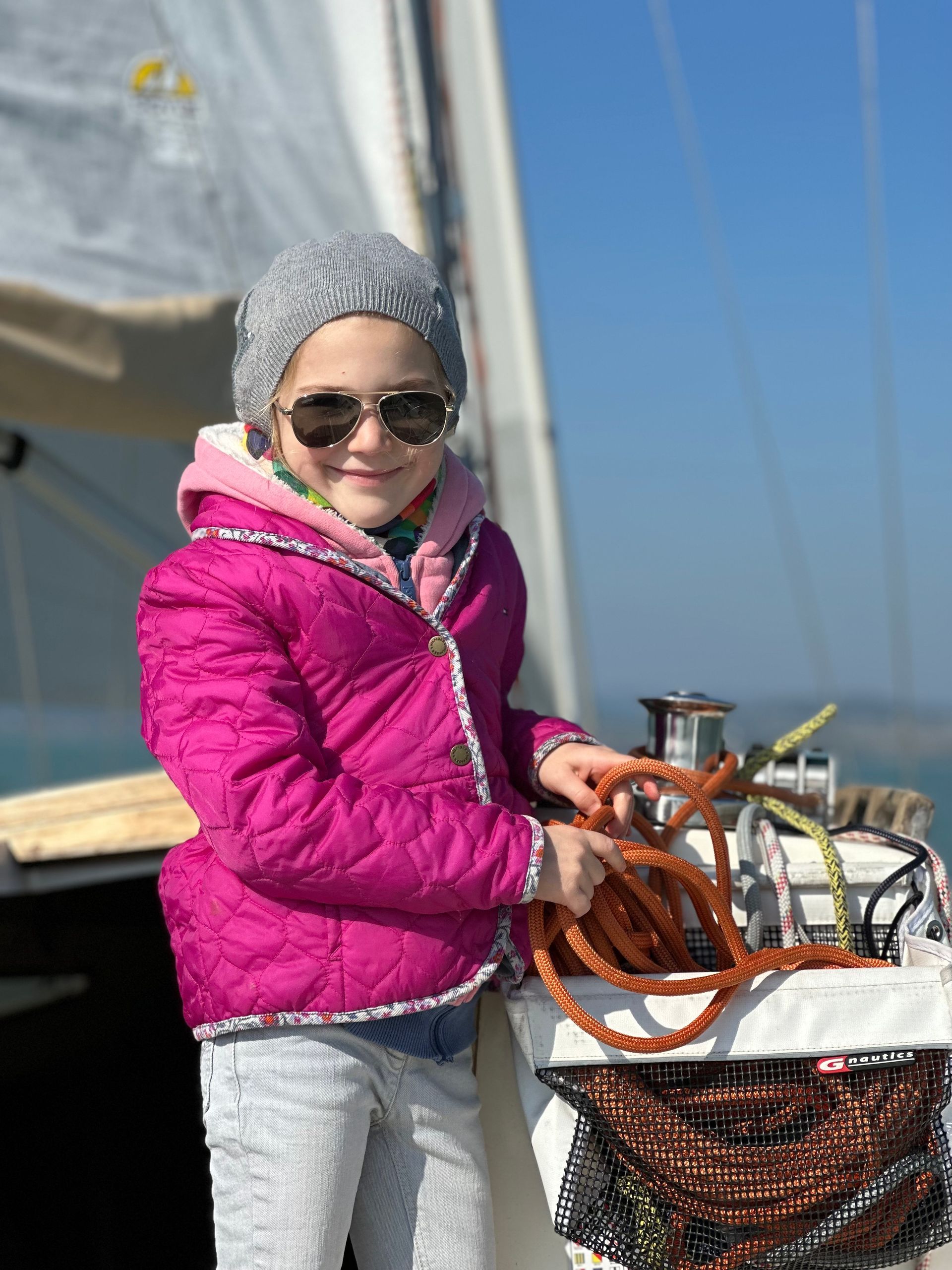 Girl on a sailboat wearing a pink jacket, sunglasses, and knit hat, holding rope, smiling.
