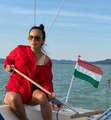 Woman on a sailboat, holding a wooden tiller, with a Hungarian flag flying.