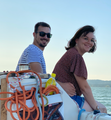 Man and woman smiling on a boat; man wears sunglasses, woman wears glasses; nautical ropes and water visible.
