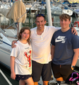 Man with two teens on a boat dock, smiling. Daylight, boats in background.