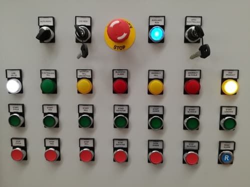 Pannello di controllo industriale con file di pulsanti etichettati, spie luminose e un interruttore di arresto di emergenza rosso.