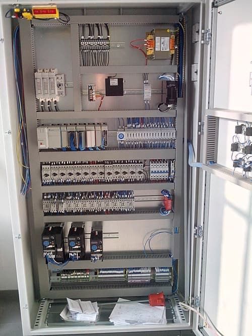 Quadro elettrico aperto con fili, interruttori, relè e sezionatori all'interno di un armadio grigio.