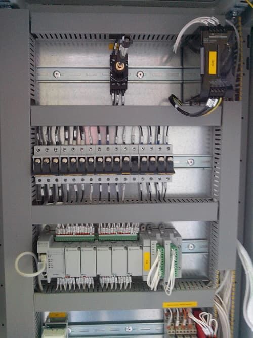 Quadro elettrico di controllo con file di interruttori automatici, relè e cablaggi all'interno di un armadio grigio.