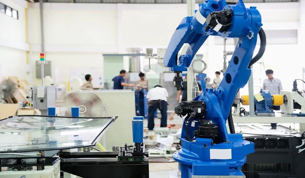 Braccio robotico industriale blu al lavoro in una fabbrica luminosa, con operai sullo sfondo.