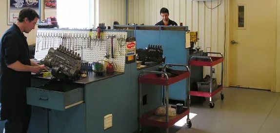 Comptoir d'atelier avec outils et fournitures ; deux personnes travaillent à des bureaux dans une petite salle industrielle.