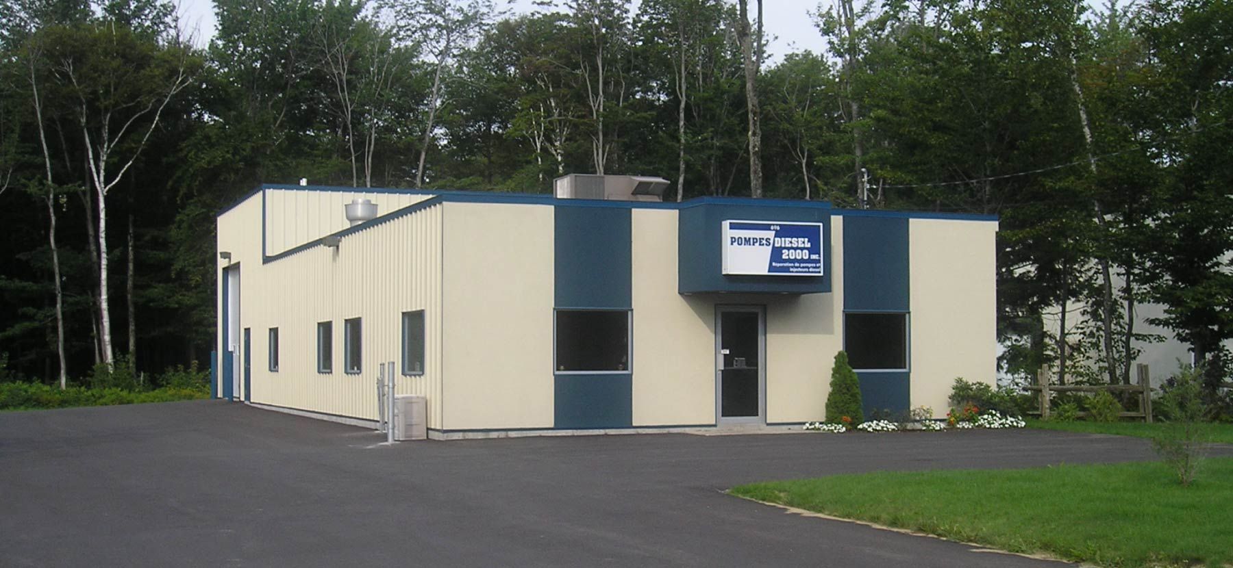 Petit bâtiment industriel beige avec des touches de bleu et une enseigne, situé à côté d'un terrain pavé et d'arbres.