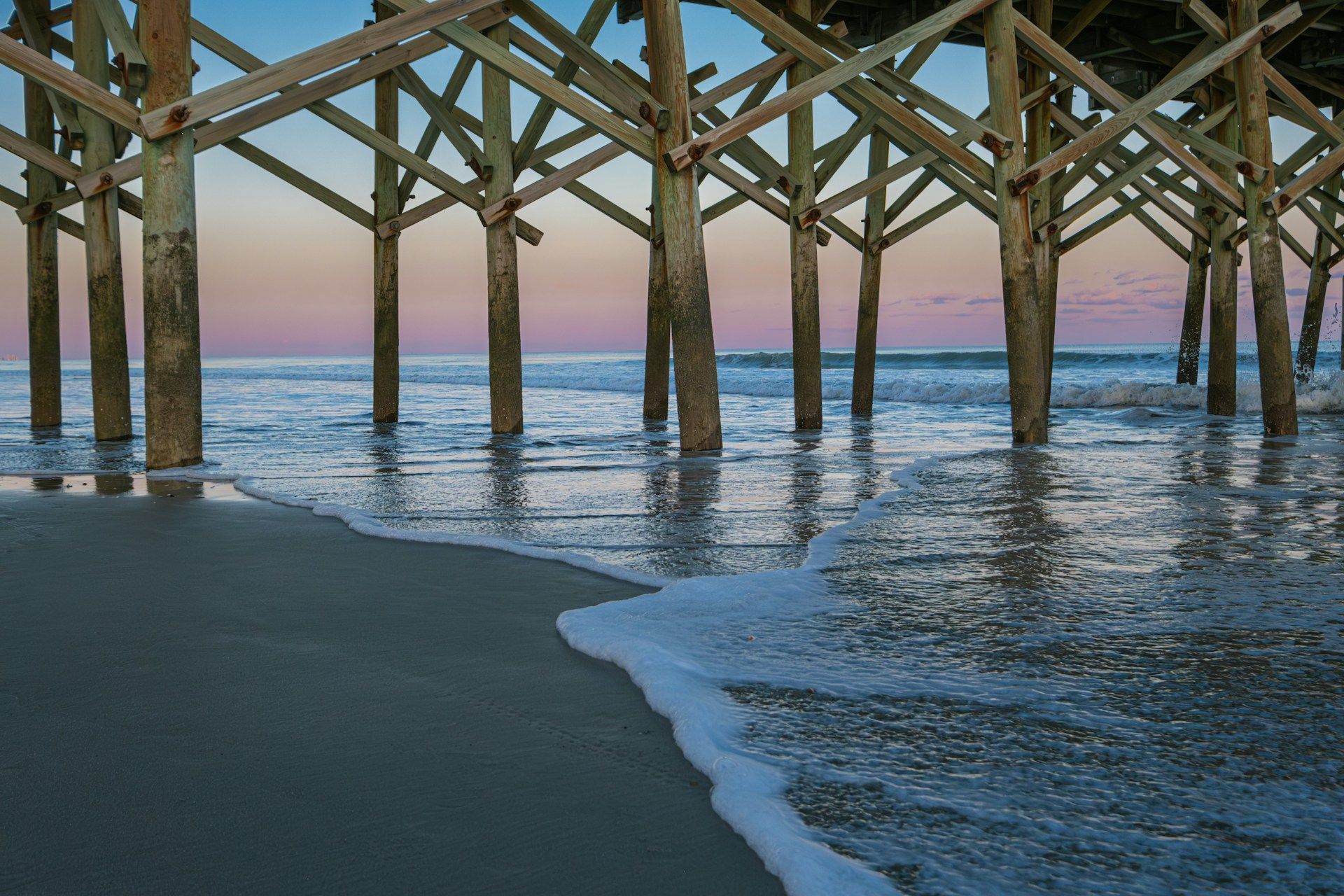 The waves are crashing on the beach under a pier.
