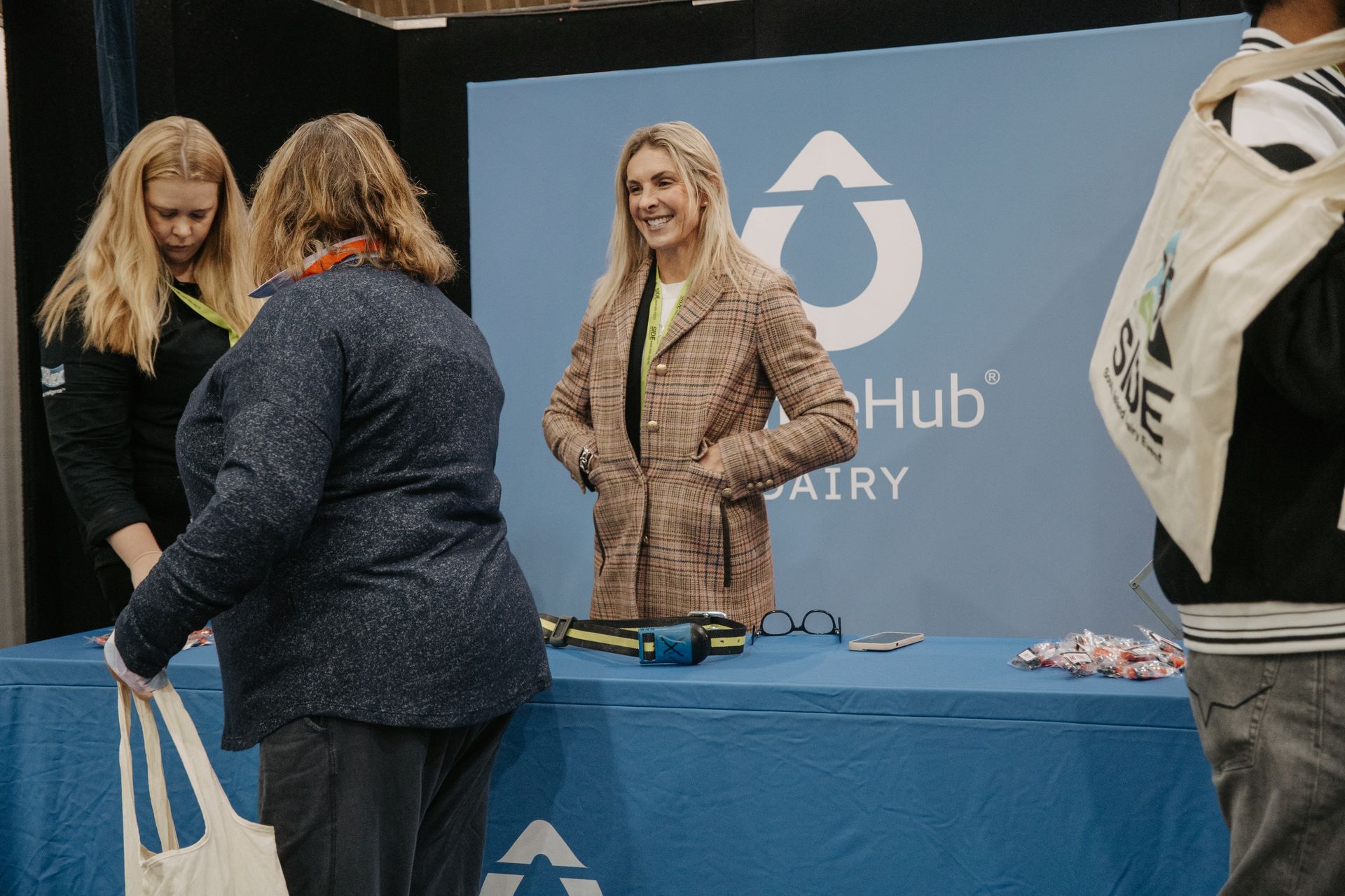 People at a trade show booth with a blue tablecloth. One woman in a tan coat smiles at a person holding a bag.
