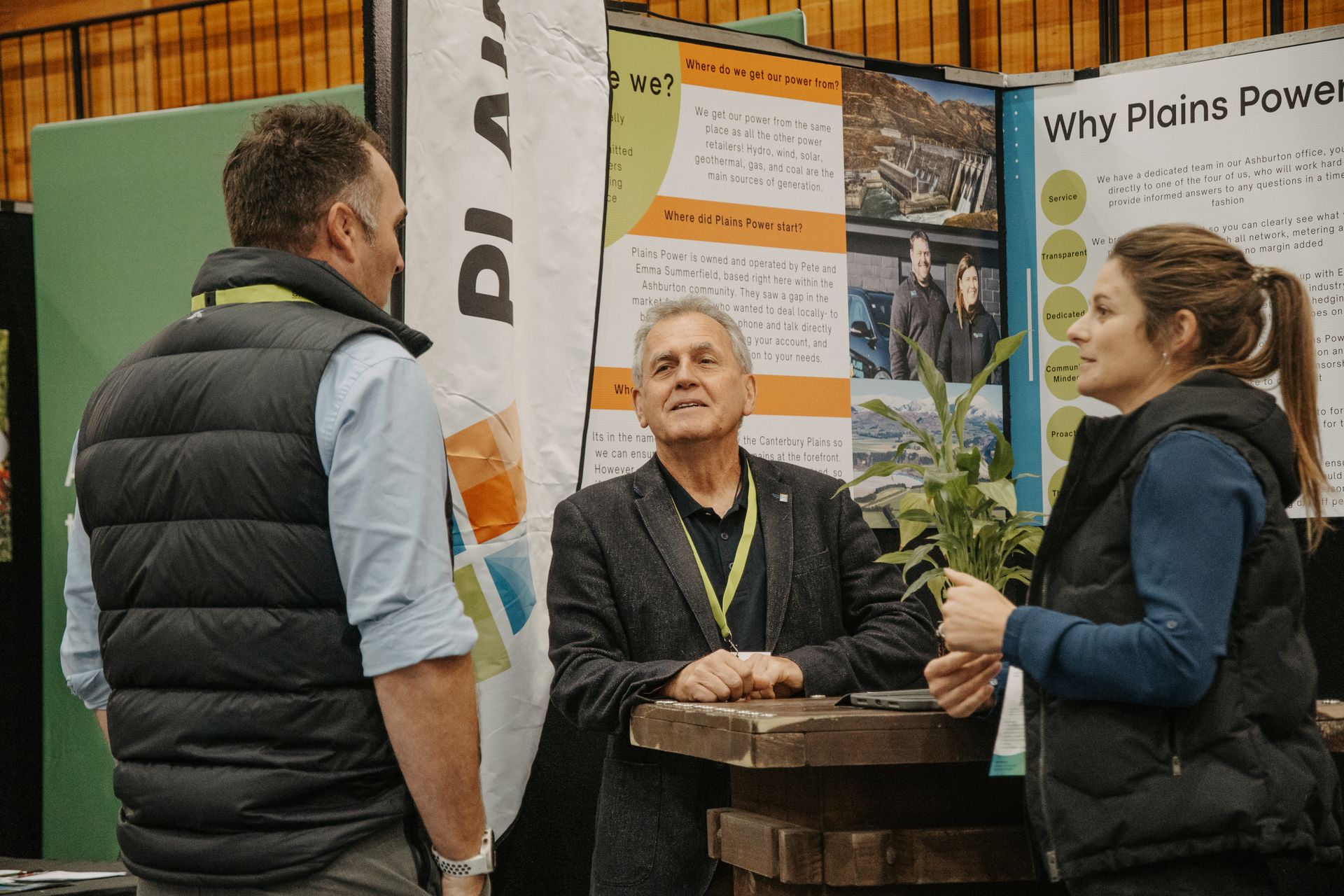 Three people at a trade show booth, discussing something. One woman holds a plant.