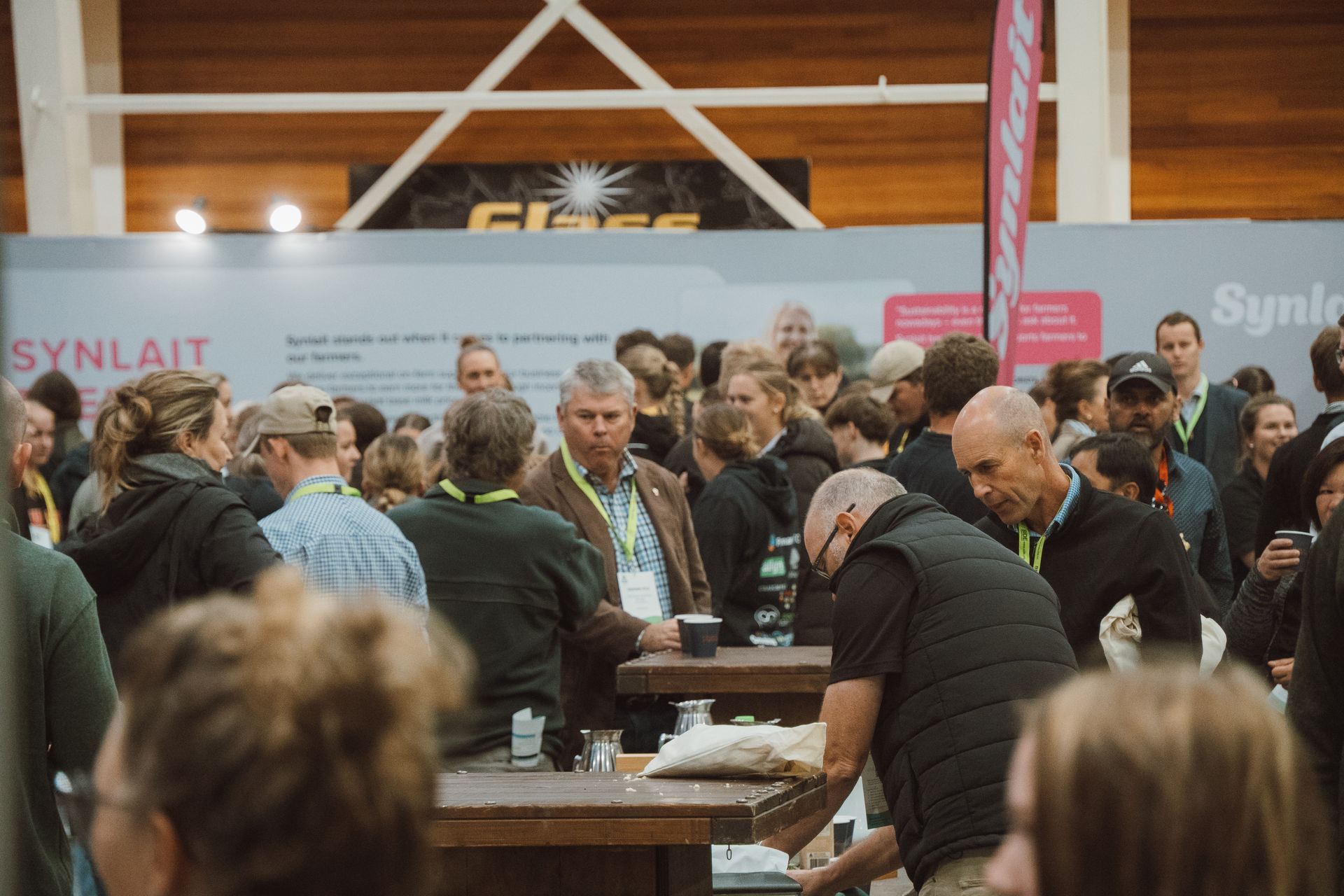 Crowd at an event, gathered around tables, interacting. Display stands in background.