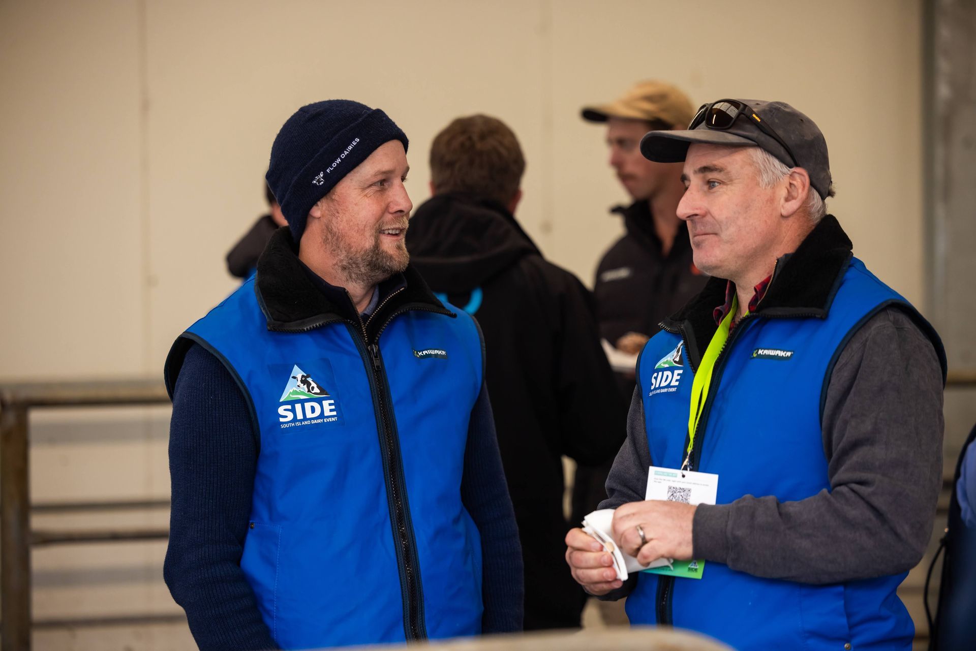 Two men in blue vests talking indoors; one wears a beanie, the other a baseball cap.
