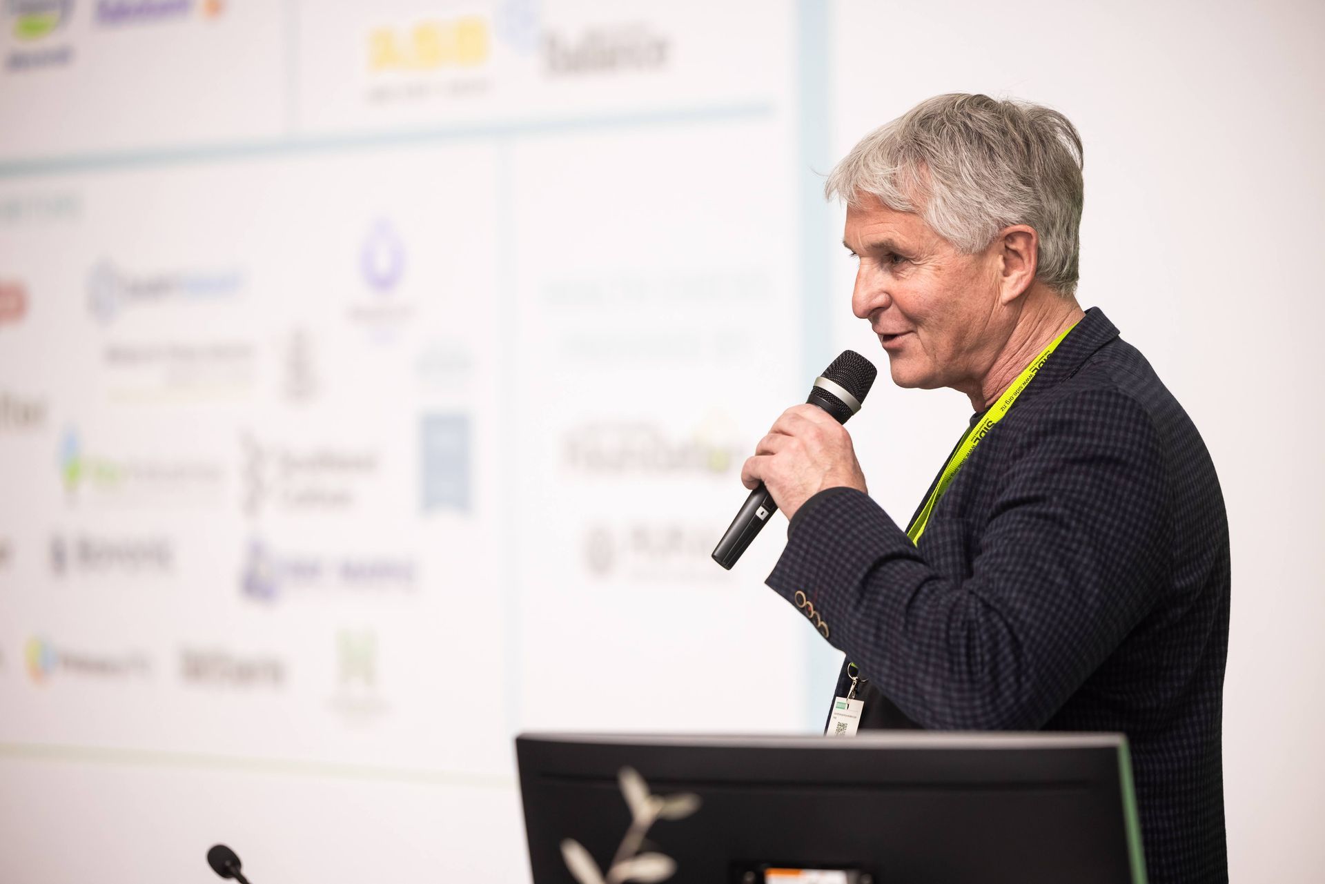 Man speaking into a microphone, standing in front of a presentation screen, wearing a dark blazer.