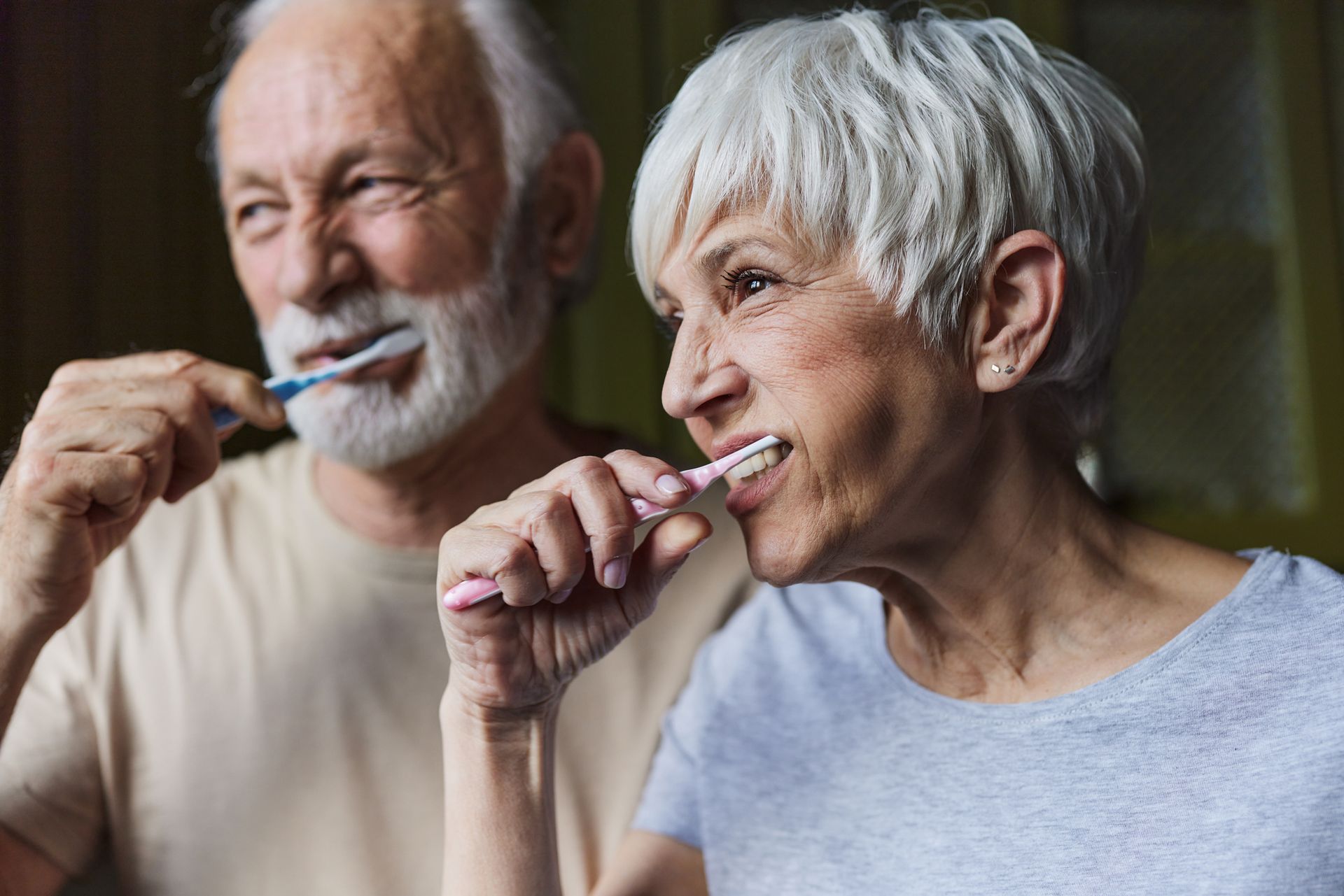 Senior couple brushing their teeth together, smiling. Light skin, gray hair, beige shirts.