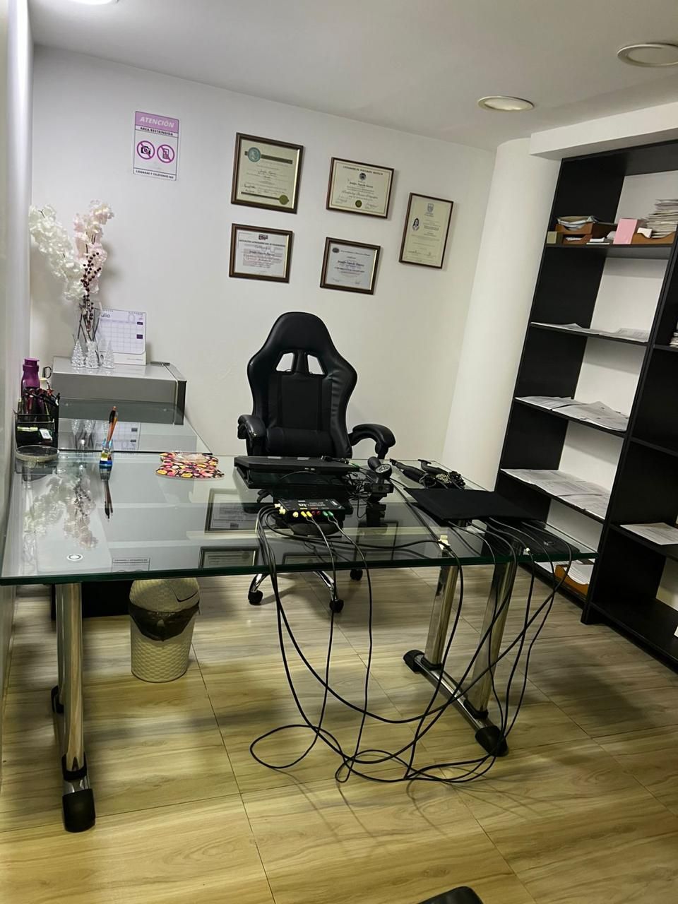 Escritorio moderno de cristal con silla negra y estantes en una pequeña oficina; los cables están a la vista en el suelo.