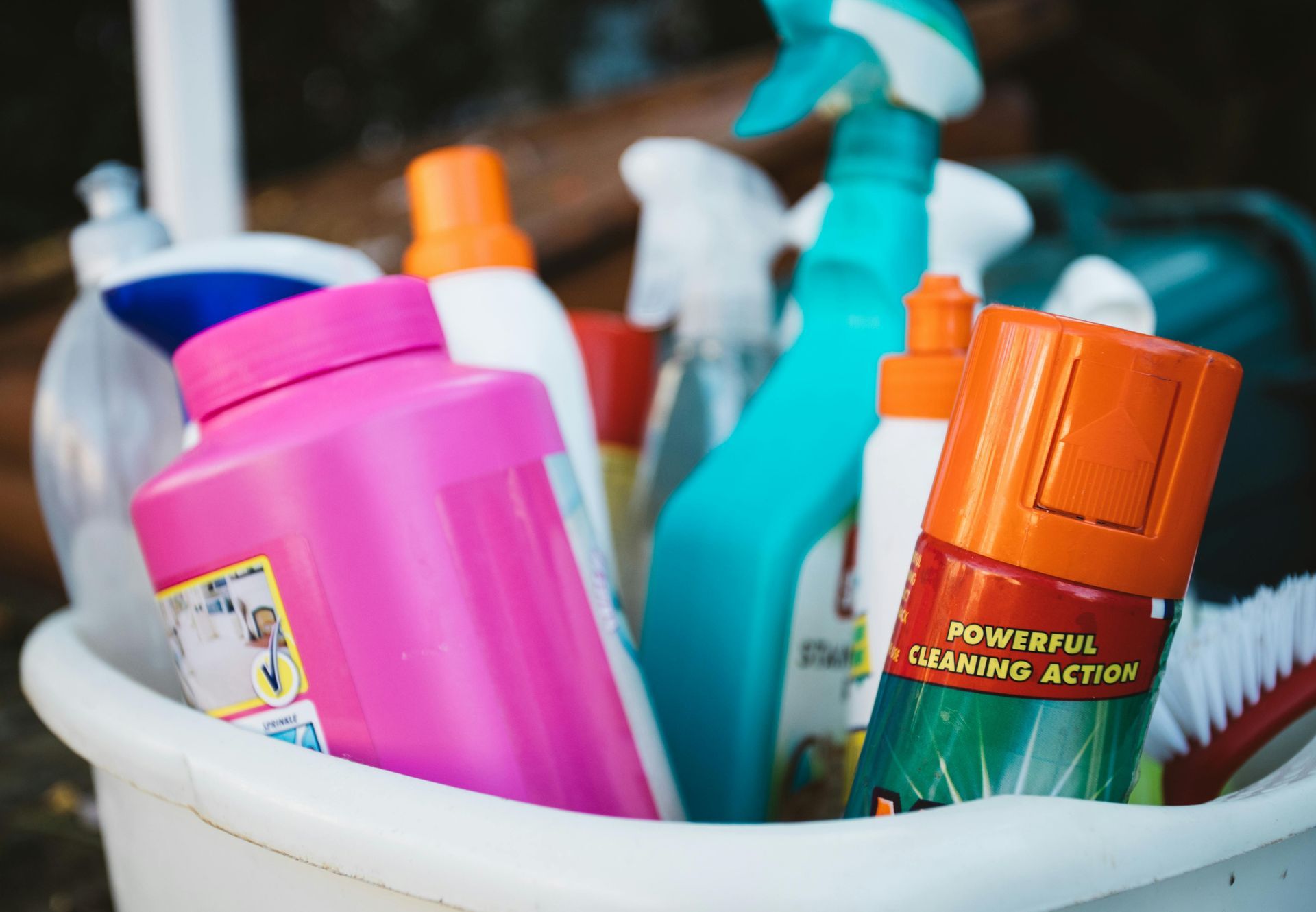Cleaning supplies in a white bucket: pink container, blue spray bottle, orange can, and brush.