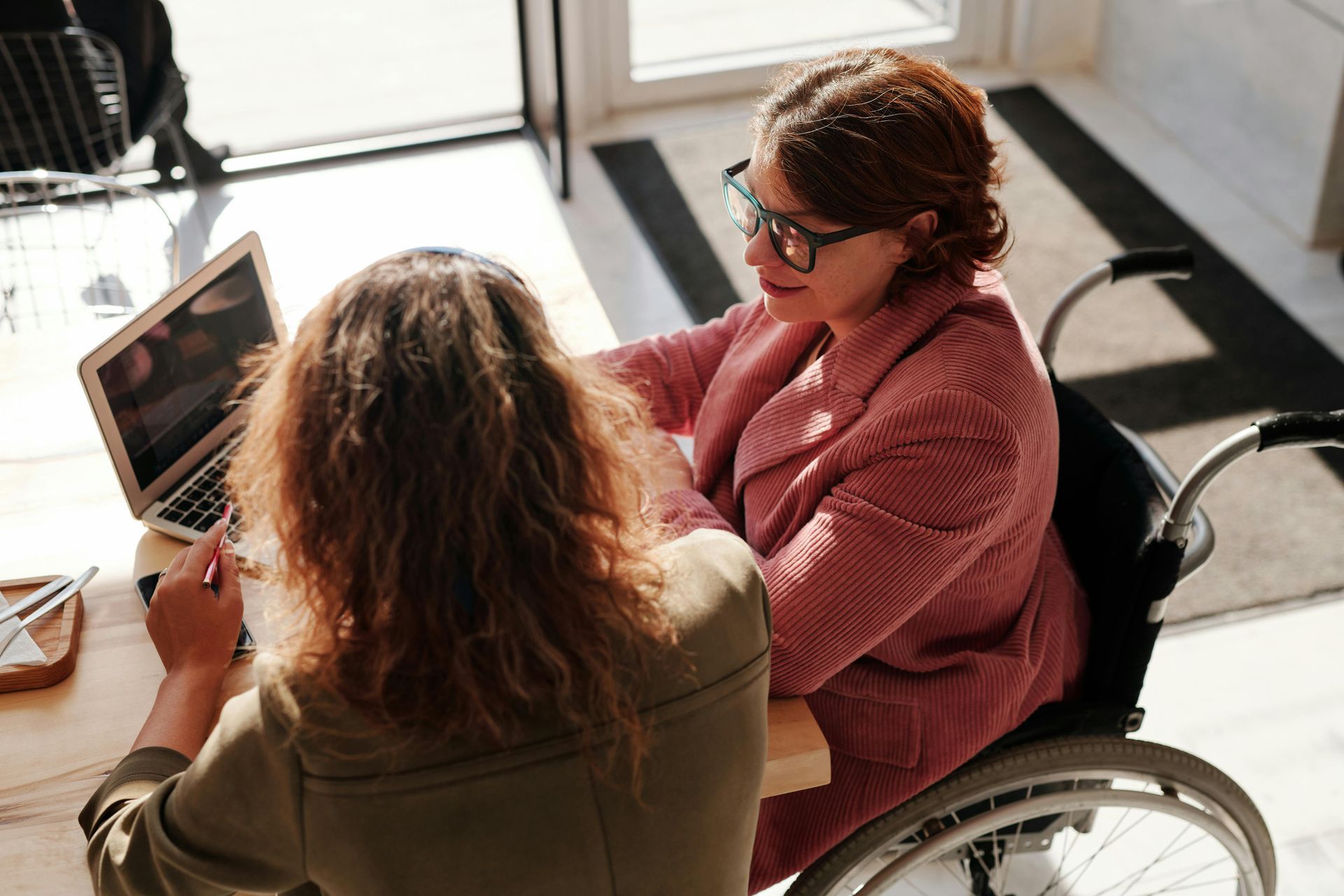 Woman in wheelchair and another woman at table with laptop, working together.