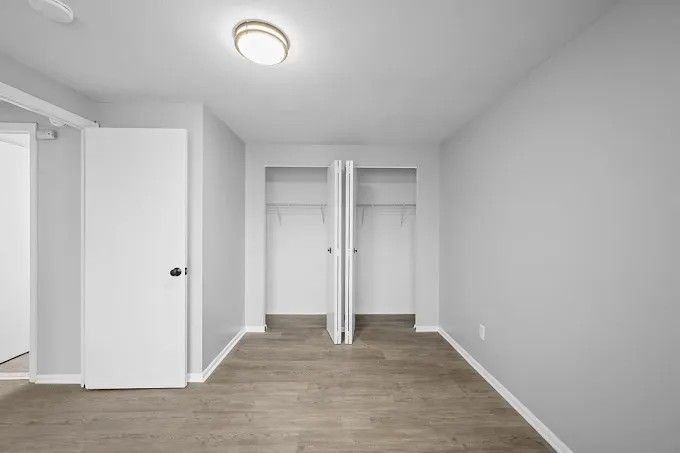 A vacant bedroom with light gray walls, brown wood-look flooring, a white door on the left, and an open double-door closet.