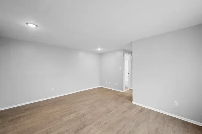A bright, empty room with light gray walls, light wood-style flooring, and recessed ceiling lights.